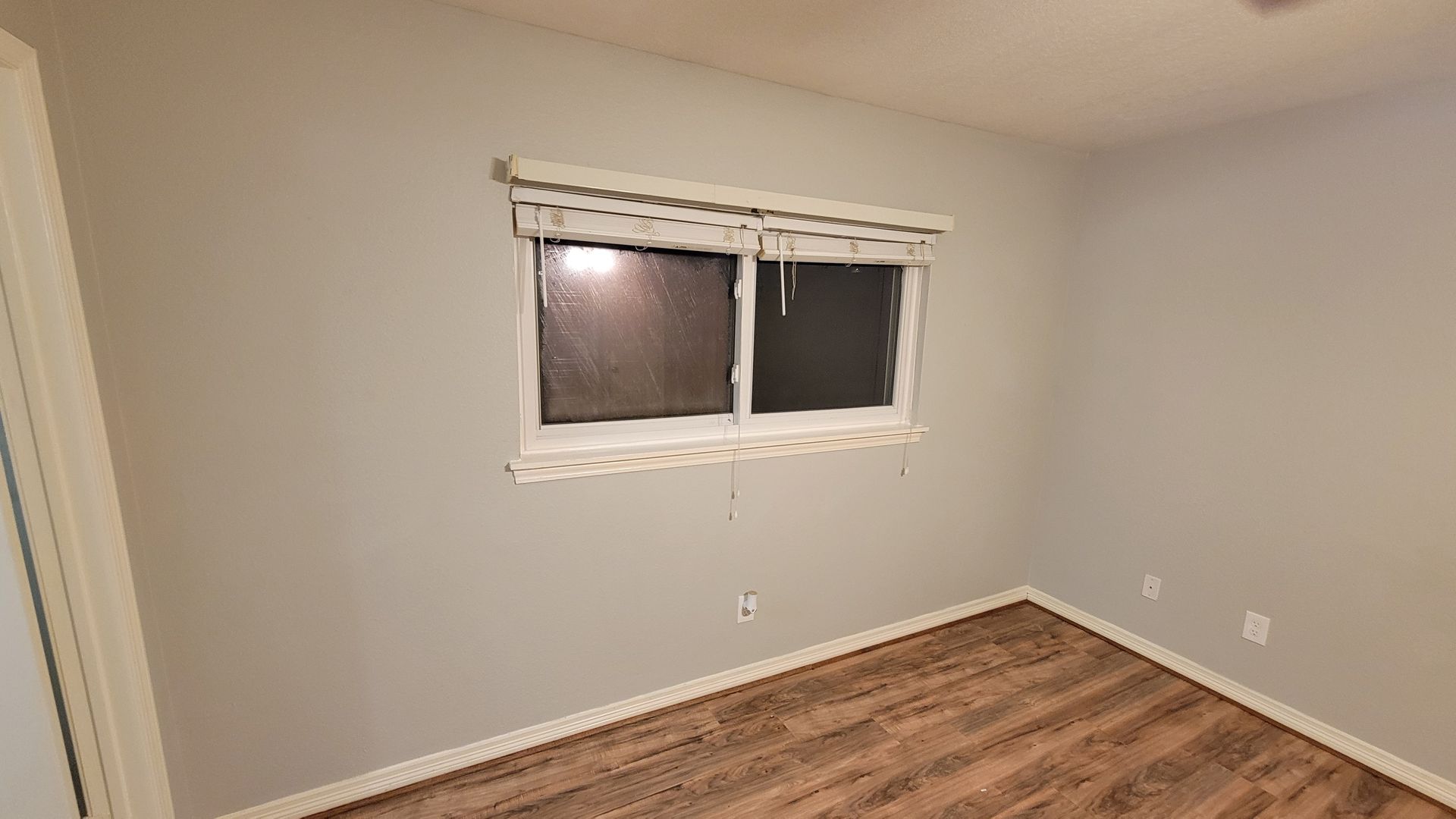 Empty room with wood-look flooring, a window with blinds, and light gray walls.