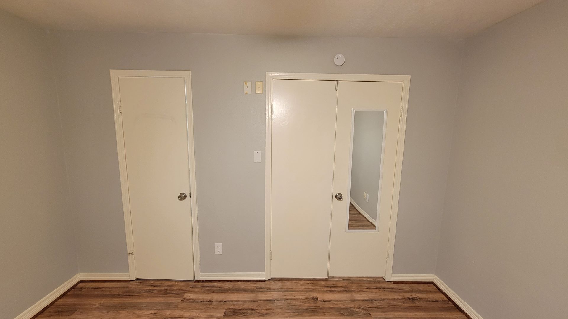 Bedroom interior with two white doors and a mirror. Gray walls, wood flooring.