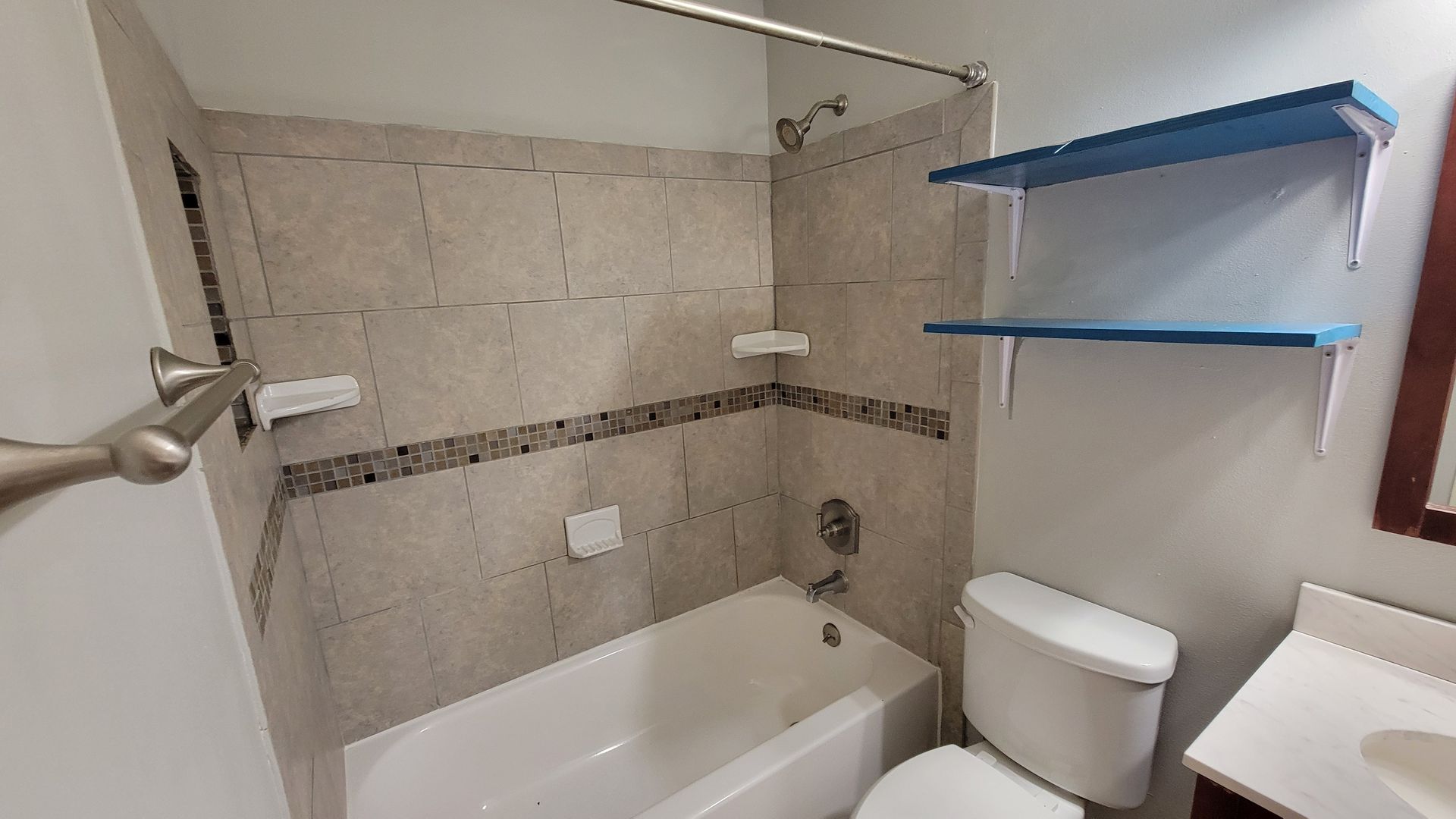 Bathroom with a white bathtub, toilet, and beige tiled walls; blue shelves.