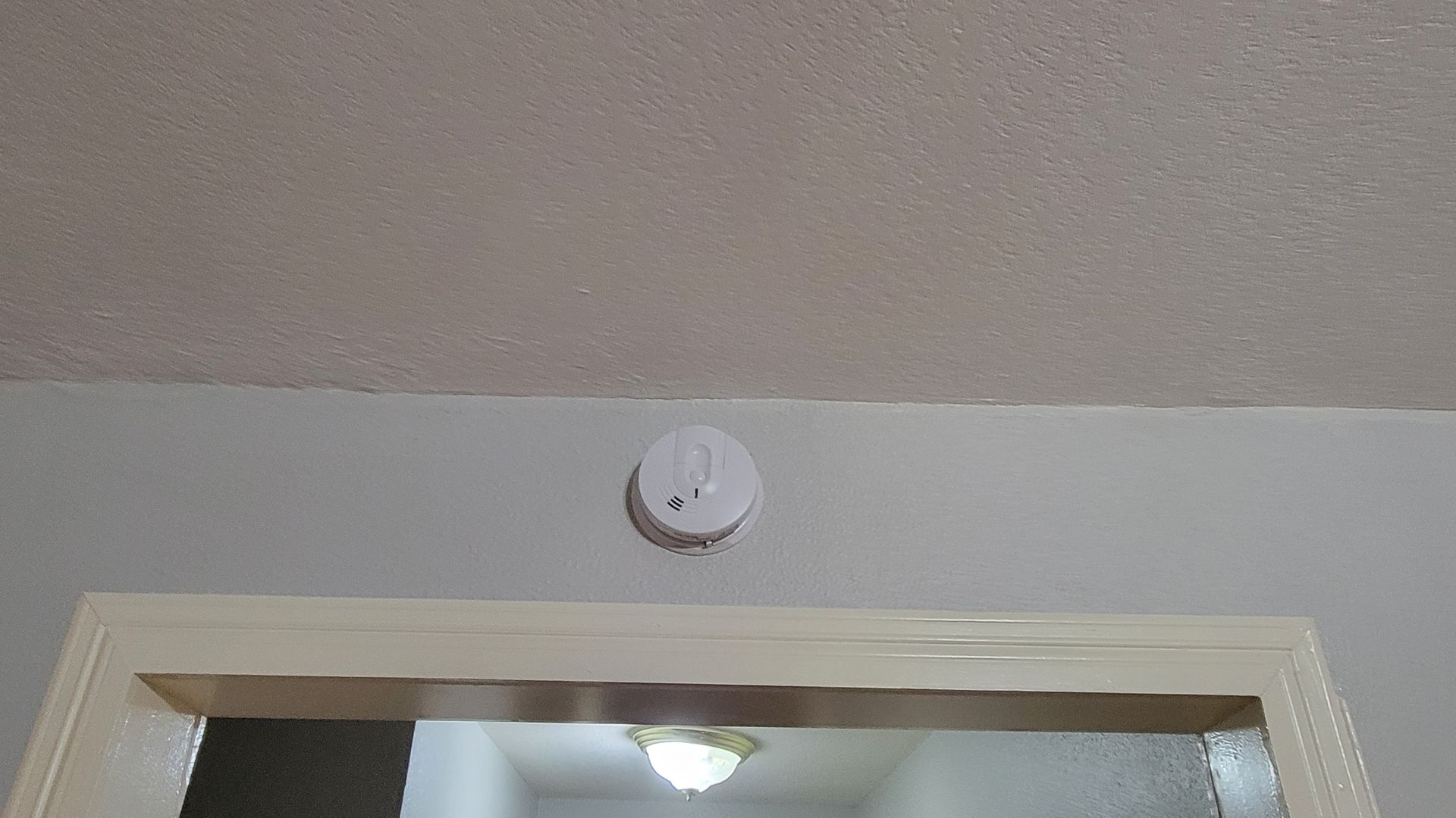 White smoke detector mounted on a white wall above a doorway with a light visible through the opening.