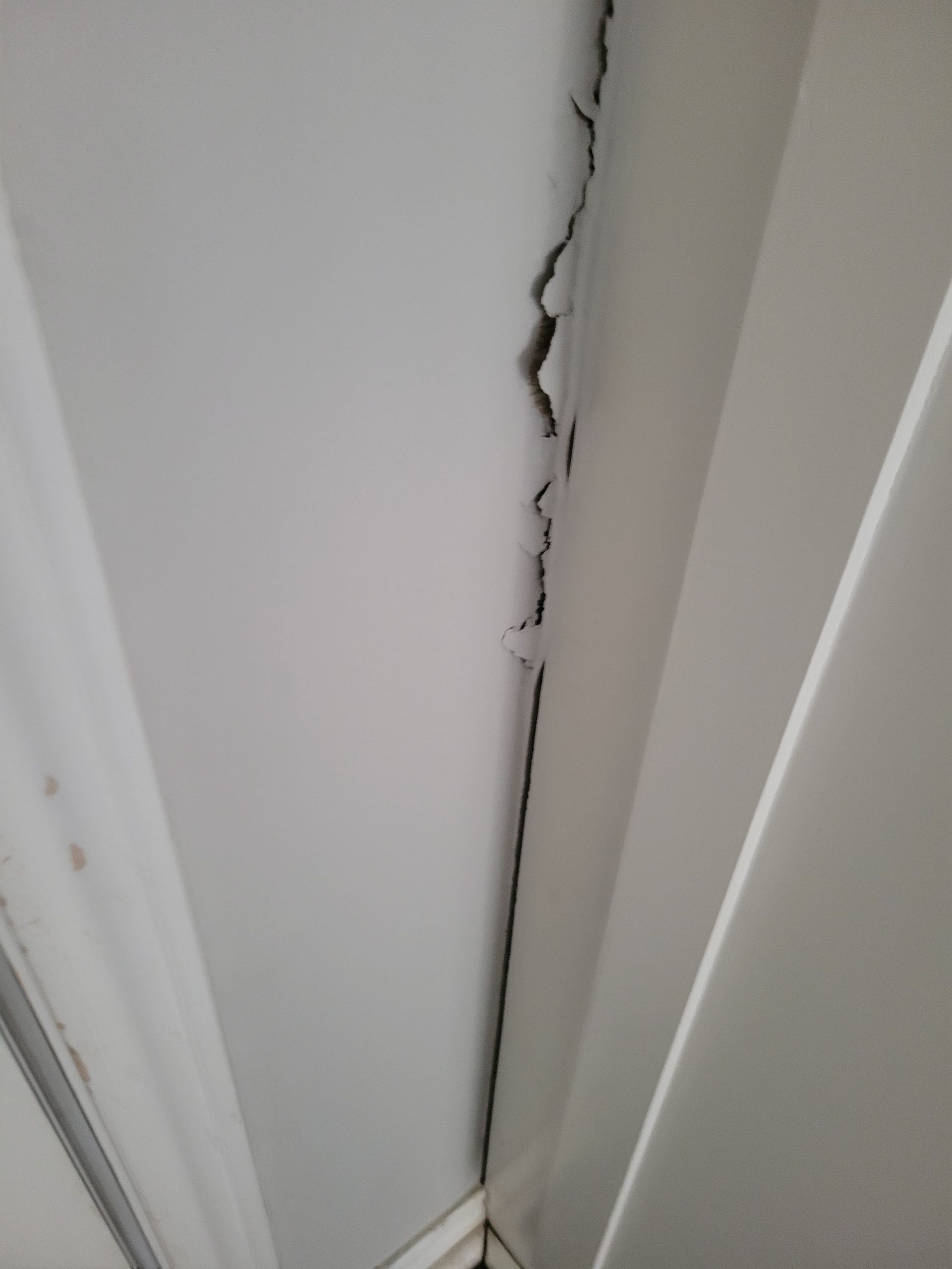 Cracked white drywall at a corner, possibly ceiling or wall, showing structural damage.