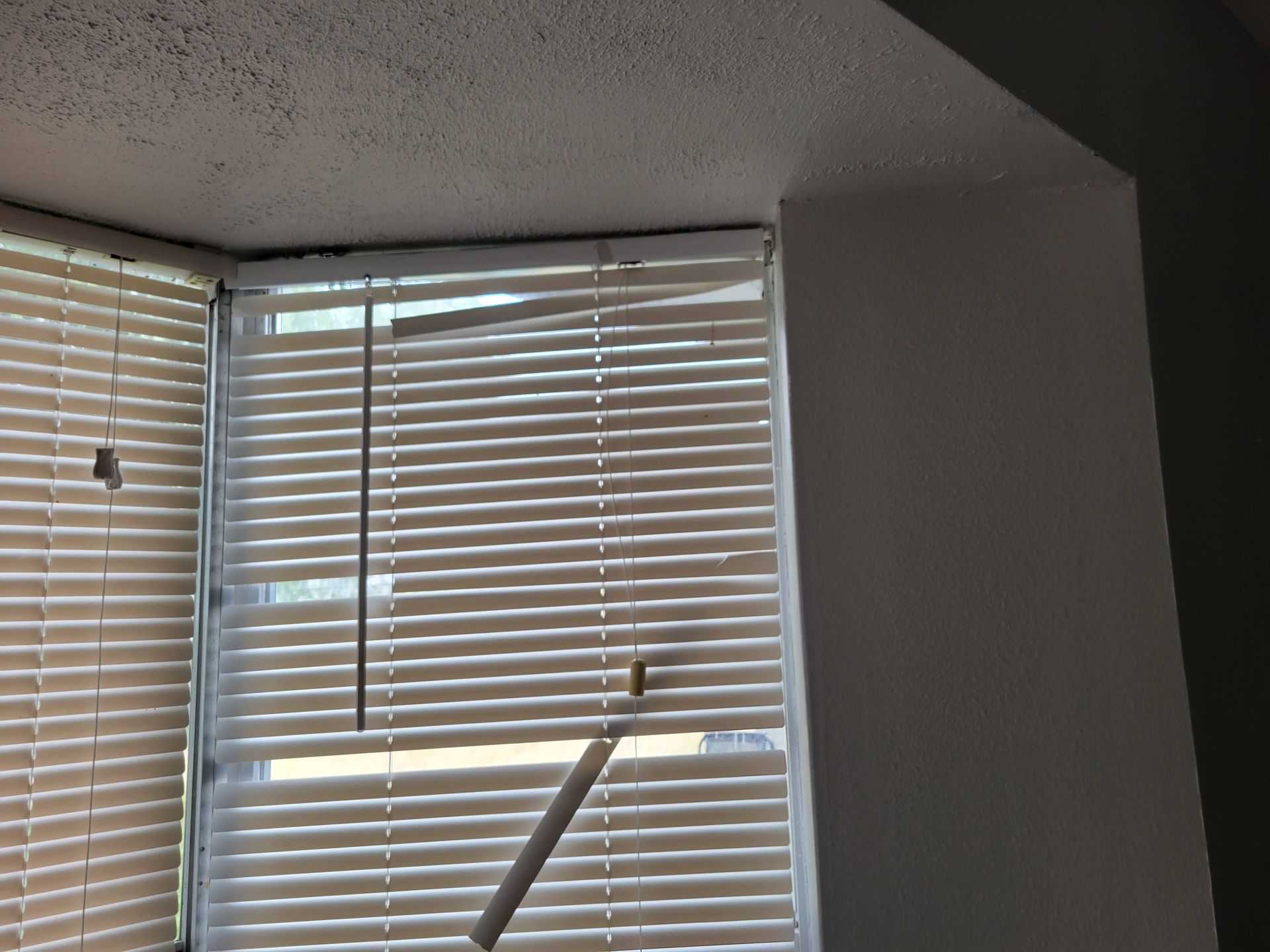 Window blinds, one panel tilted open, in a corner window. Beige blinds with a white wall.