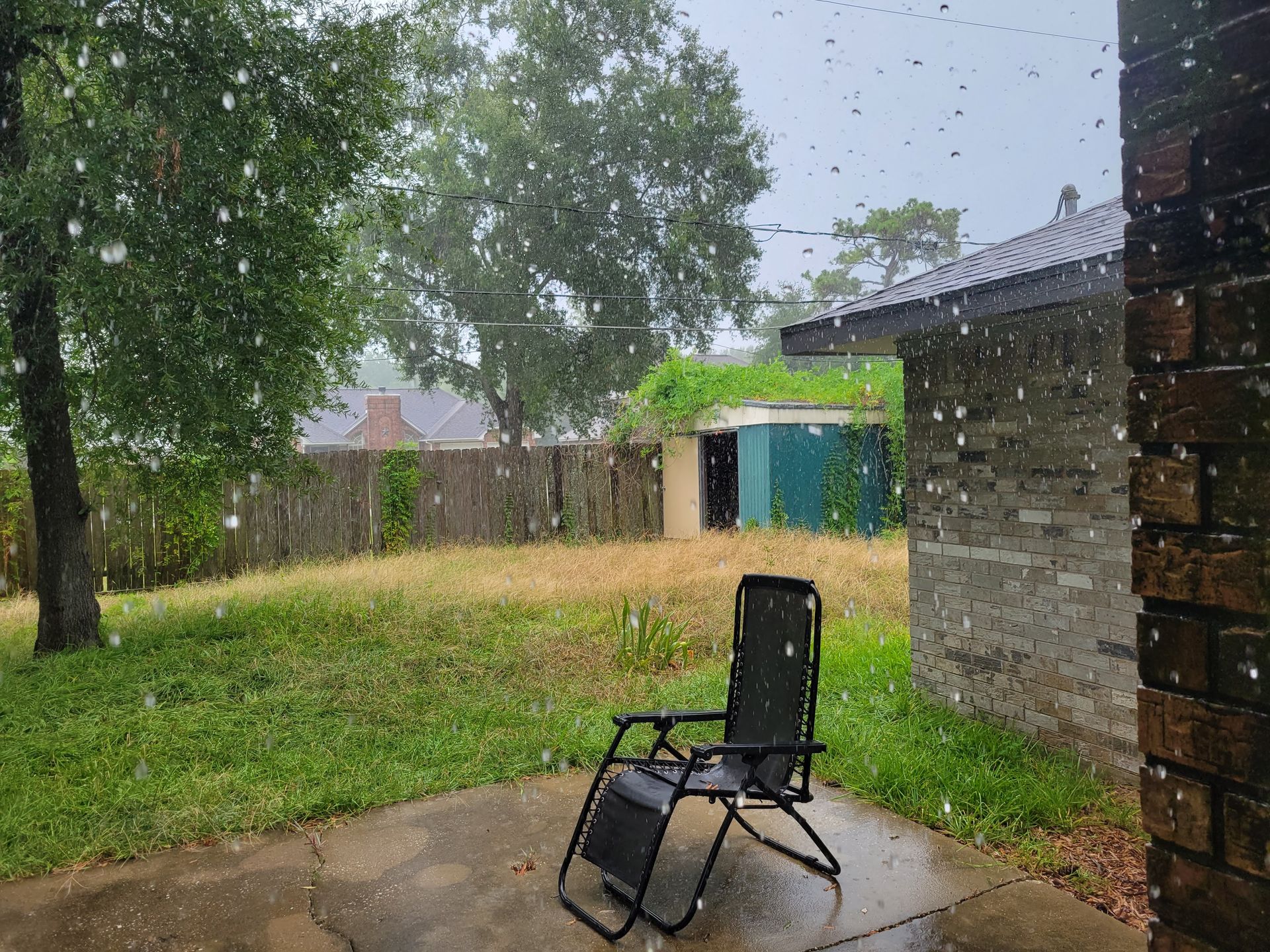 Rainy backyard scene: lawn chair on patio, rain falling, trees, and buildings visible.