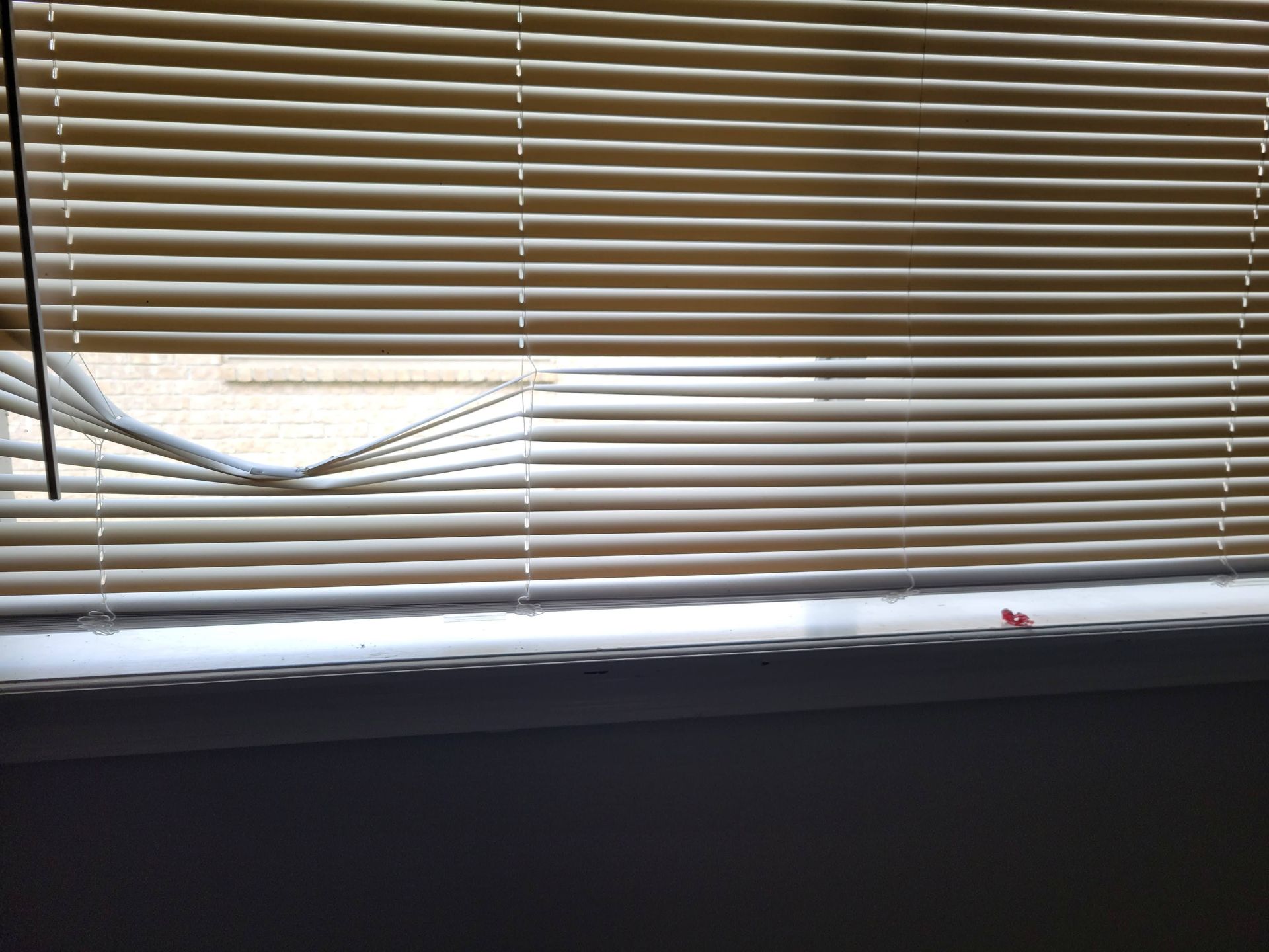 Tan blinds partially open with a broken section, revealing a window and sill.