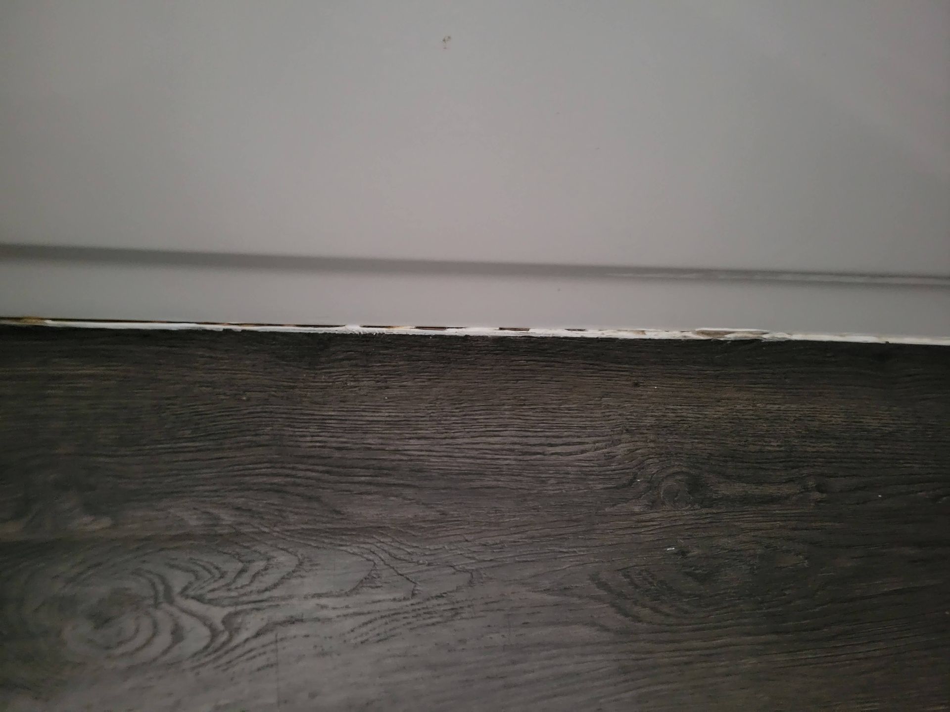 Dark wood-look flooring meets a white wall. The baseboard is visible with a slight gap.