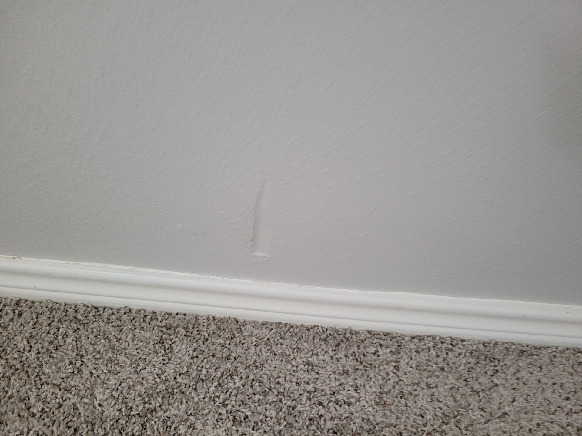 White wall with a vertical crack above a white baseboard, set over a gray carpet.