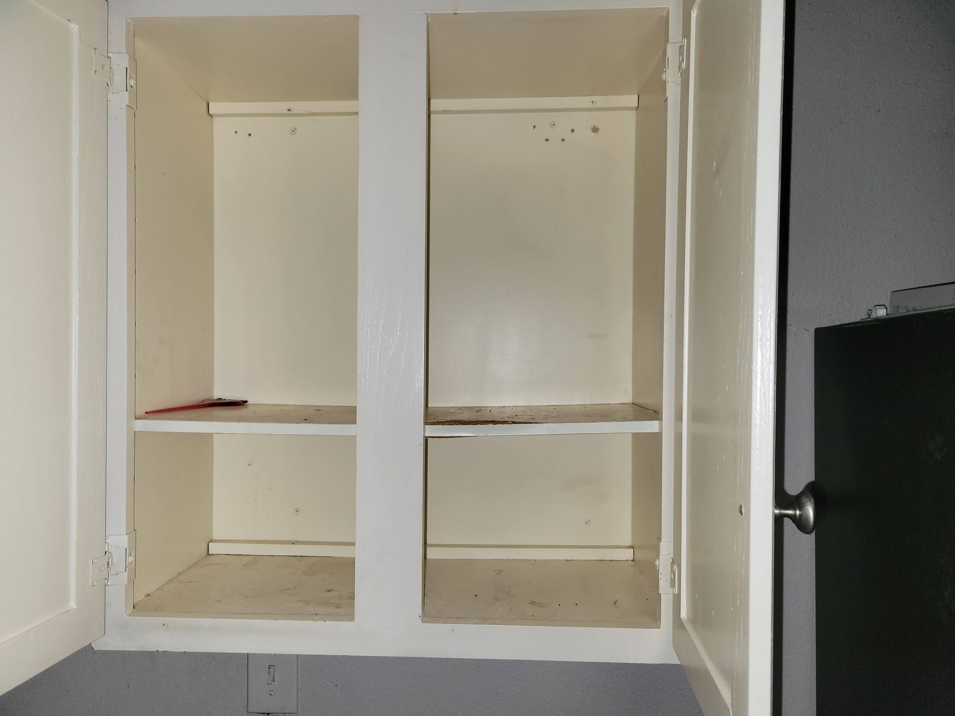 Empty, open white kitchen cabinet with two shelves. Beige interior, white frame. Dark wall in the background.