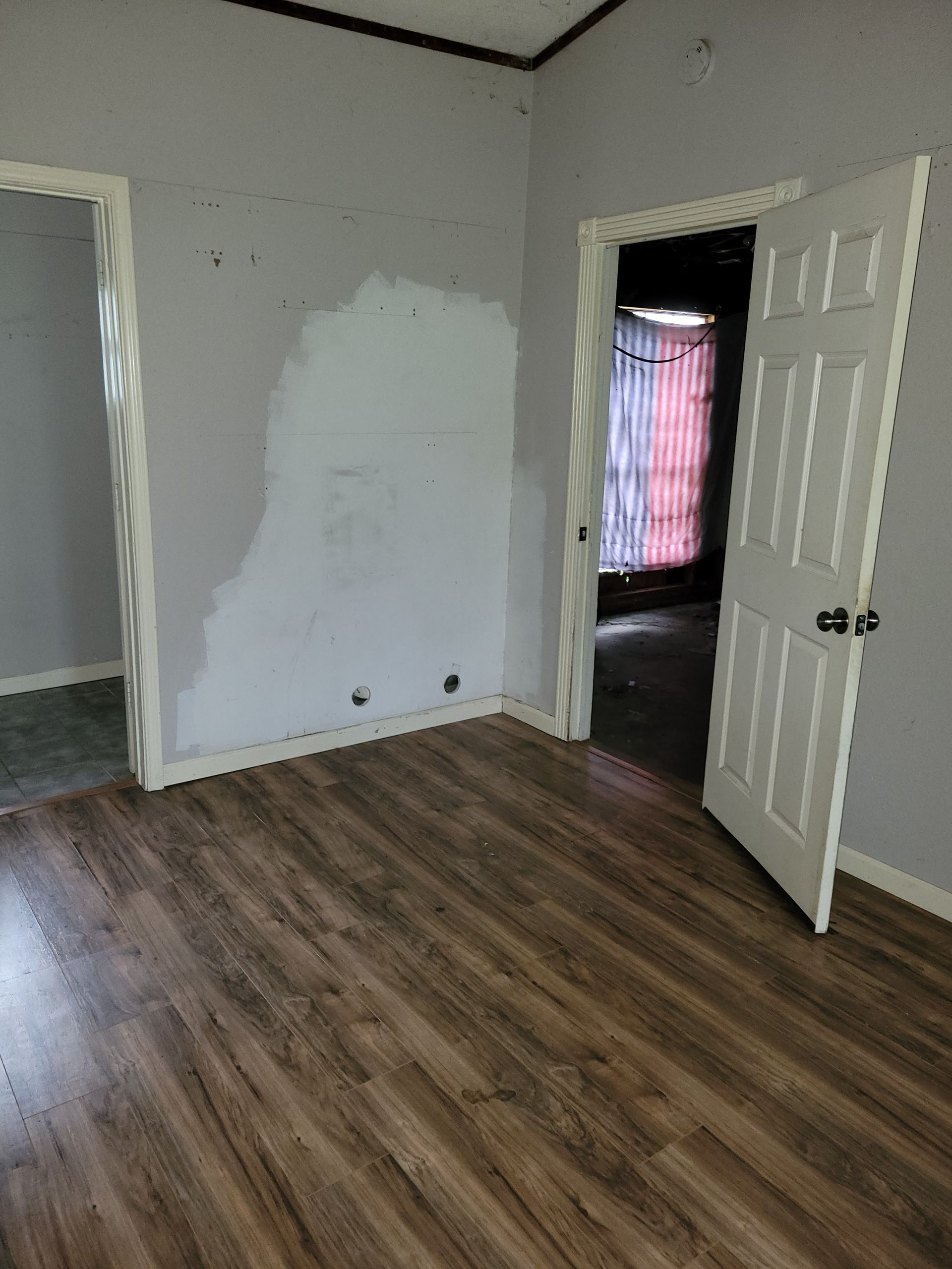 Room with wood-look floor, gray walls, two doorways, and a partially open white door. One wall has a patched area.