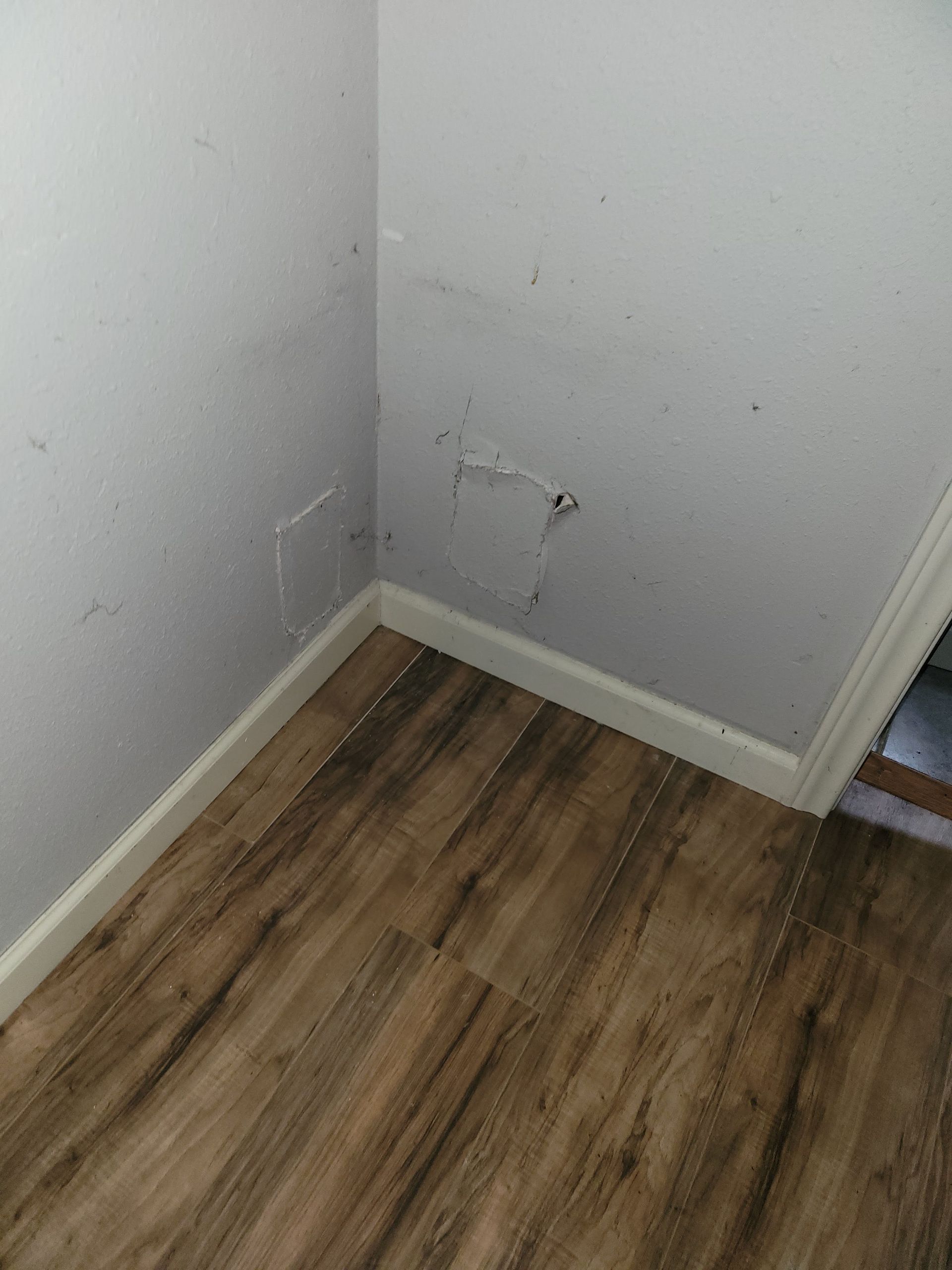 Corner of a room with light wood-look flooring and white walls. A damaged square area is visible on one wall.