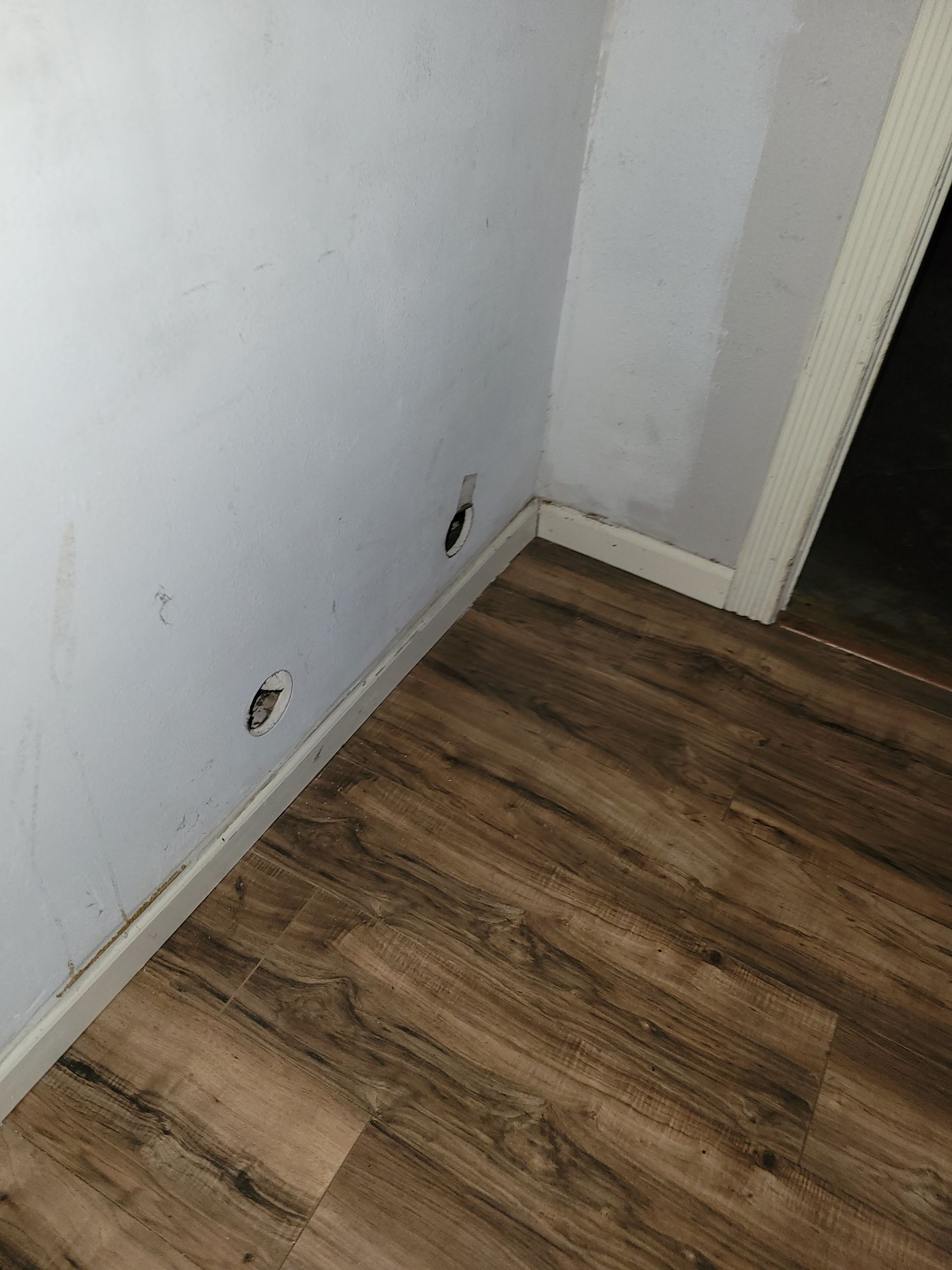 Two holes in a light-colored wall, near a wood-look floor and baseboard. A door frame is visible.