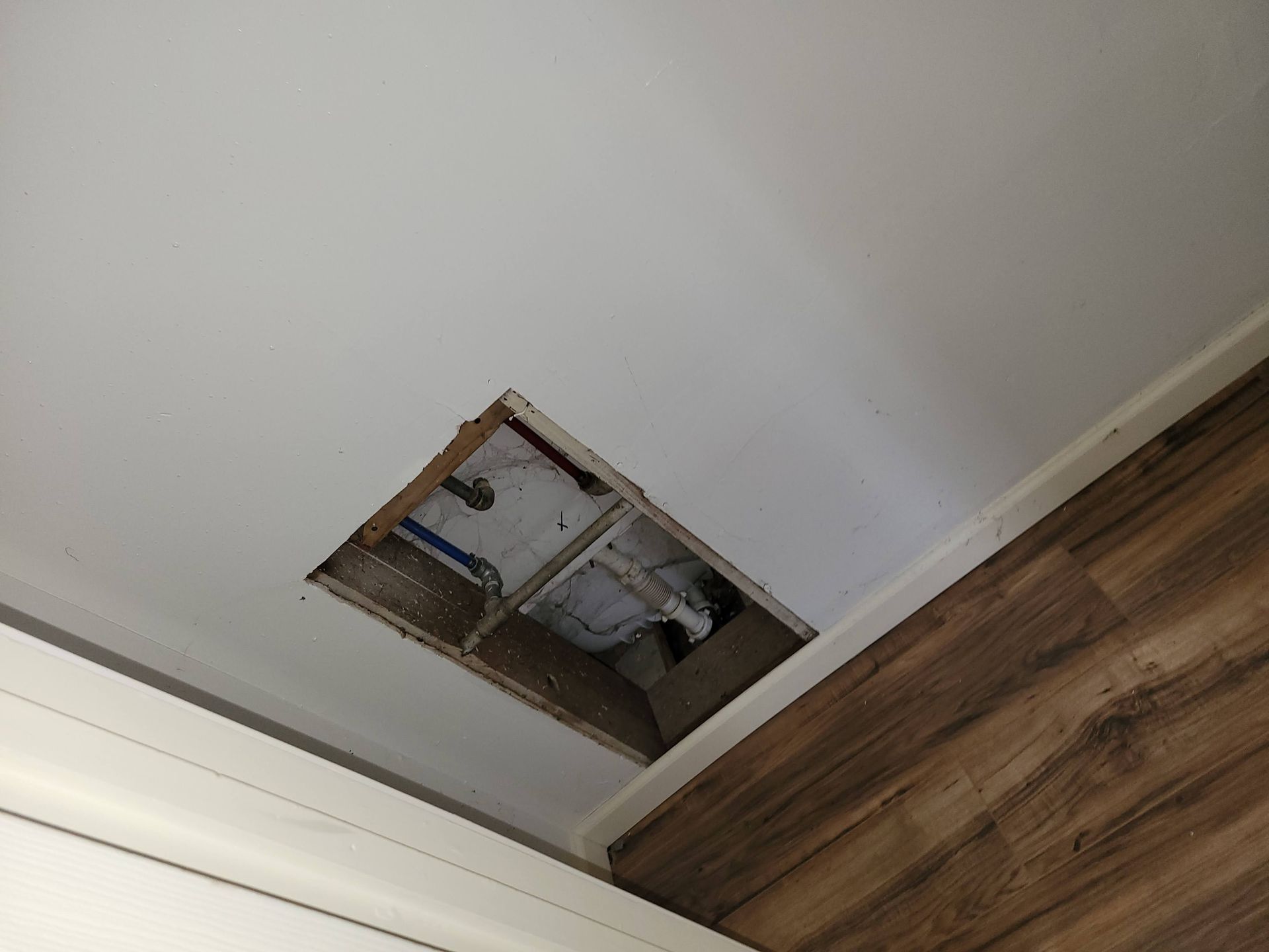 Hole in white ceiling, showing wooden frame and plumbing; corner of room with brown flooring and white trim.