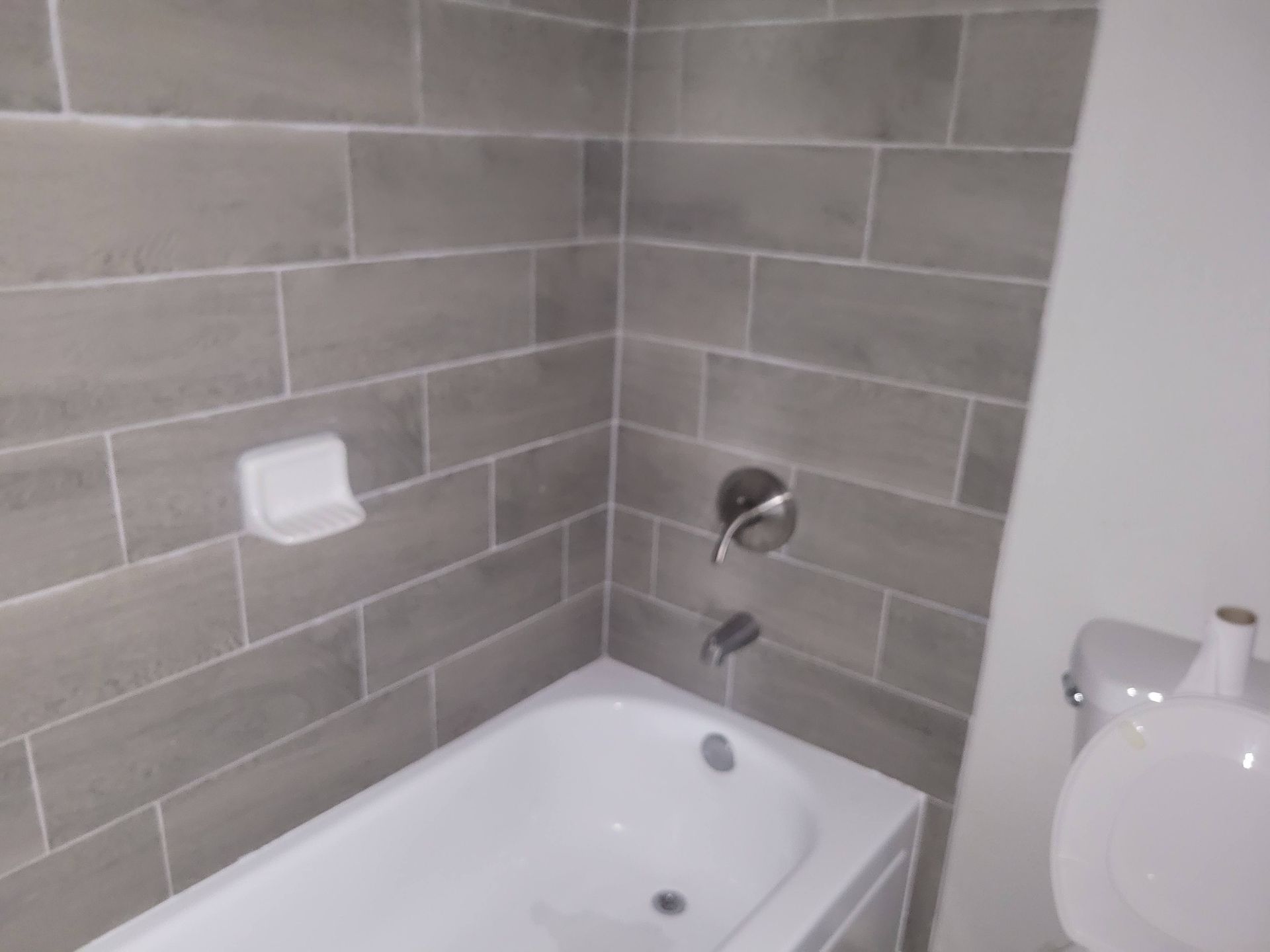 Gray-tiled bathroom with a white tub and toilet, soap dish, and faucet.