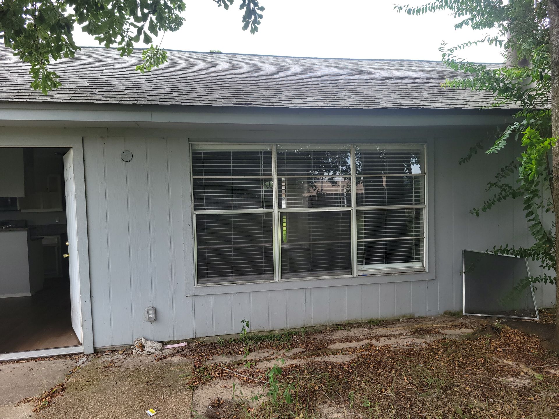 Exterior of a light blue house with a window, blinds, and a damaged roof. Debris covers the ground.