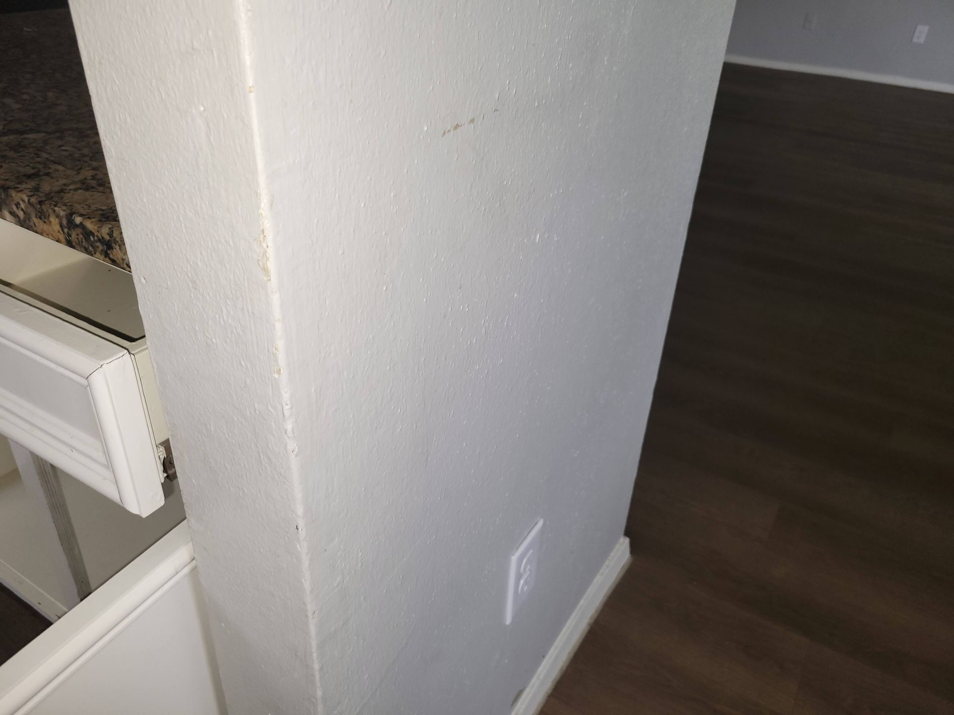 White textured wall with an electrical outlet, next to a cabinet with a countertop and dark wood flooring.