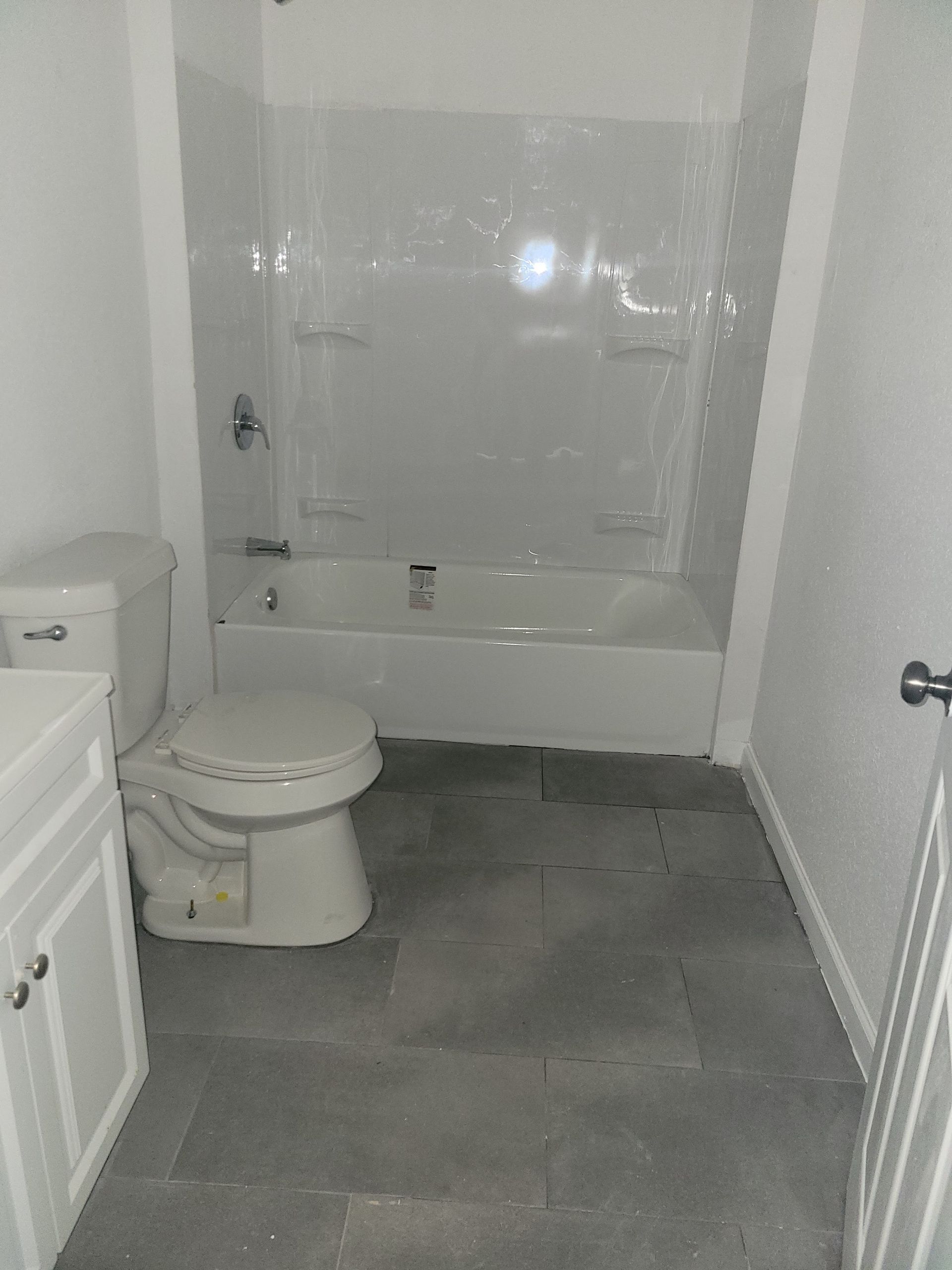 Bathroom with a white tub, toilet, and vanity against gray tiled floor and white walls.