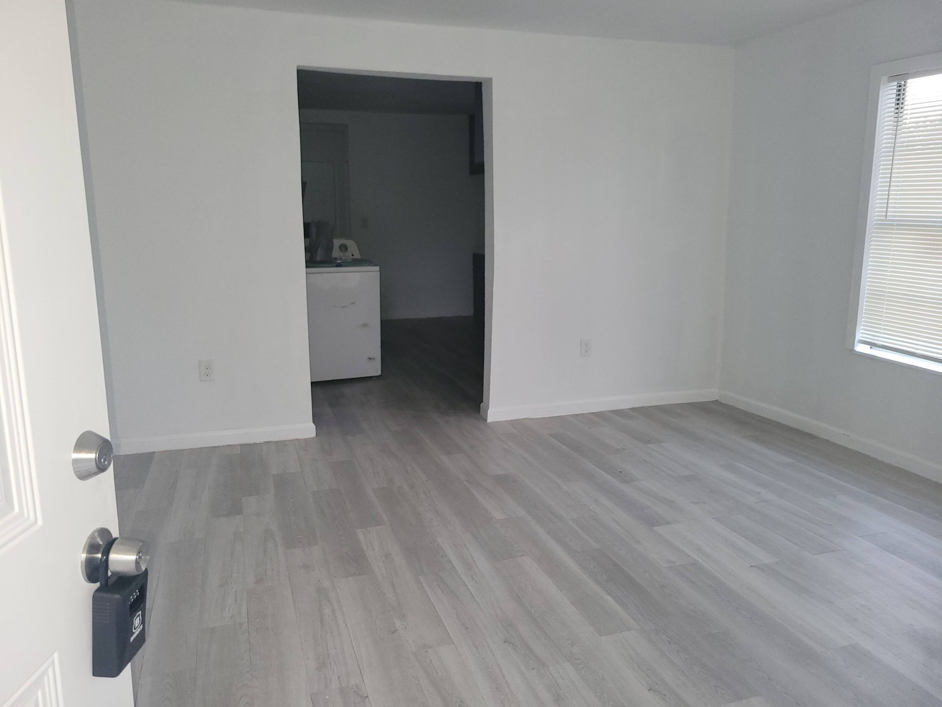 Empty room with grey wood-look flooring, white walls, and an open doorway leading to another room.
