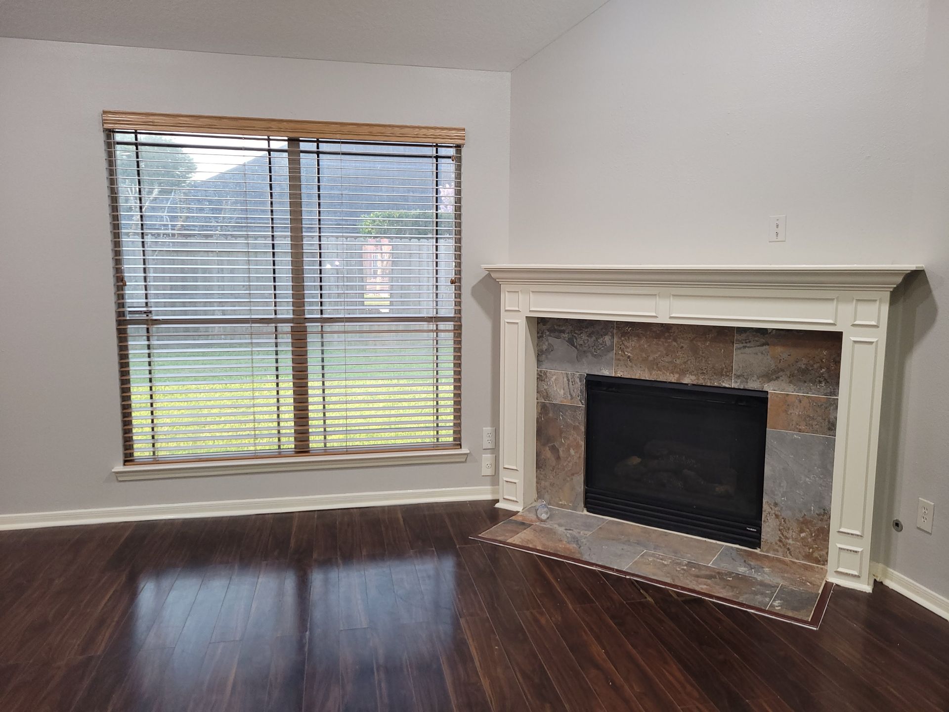 Living room with a fireplace, window with blinds, and wood flooring.