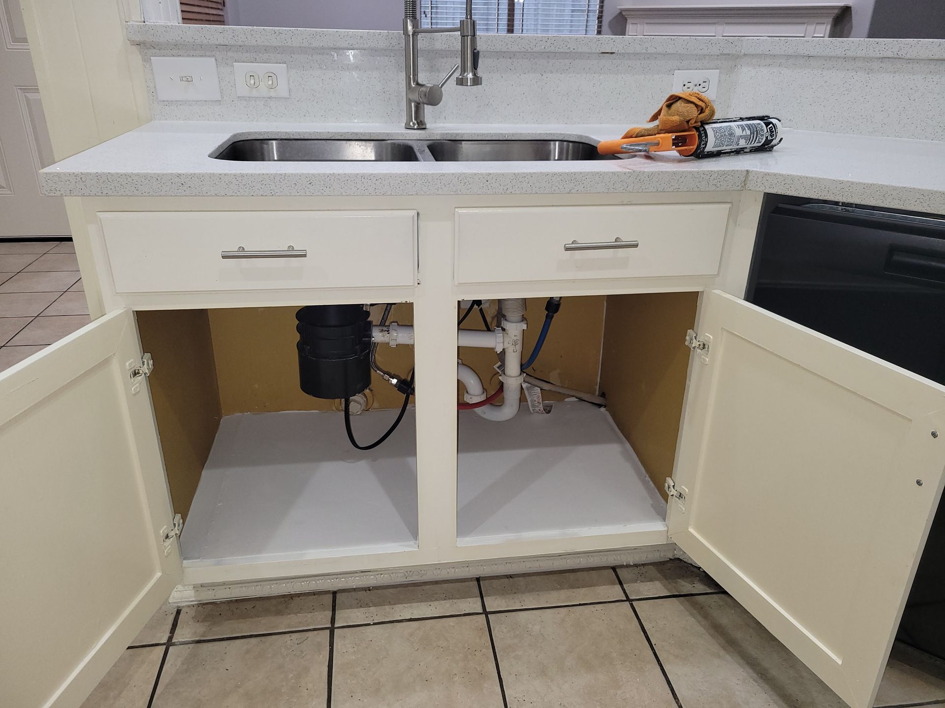 Kitchen cabinet open, showing sink plumbing, a garbage disposal unit, and light interior.