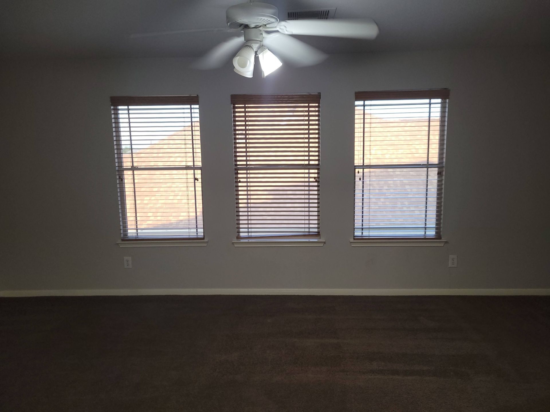 Room with three windows covered by blinds, ceiling fan, and brown carpet.