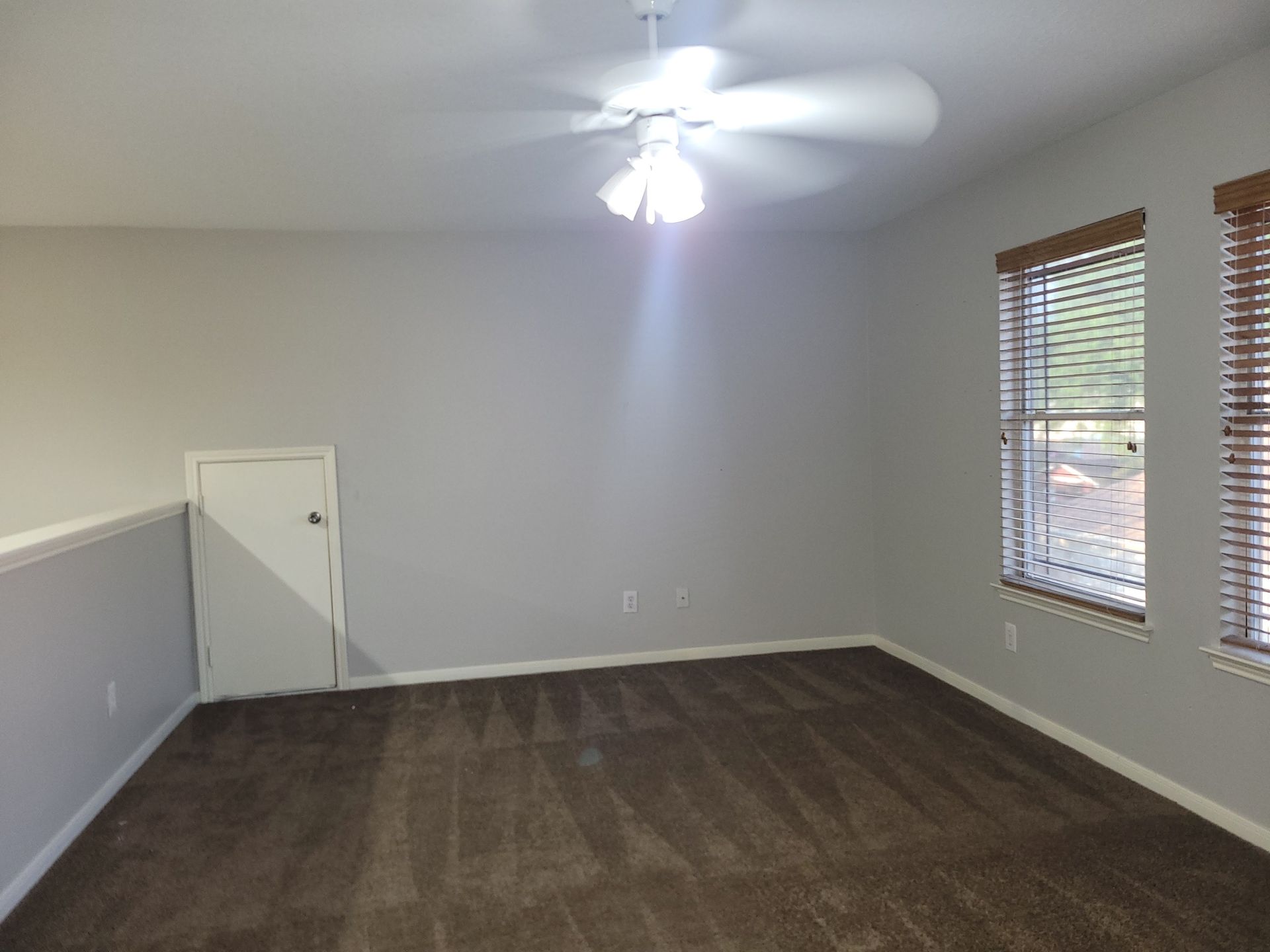 Empty room with brown carpet, gray walls, ceiling fan, small door, and window with blinds.