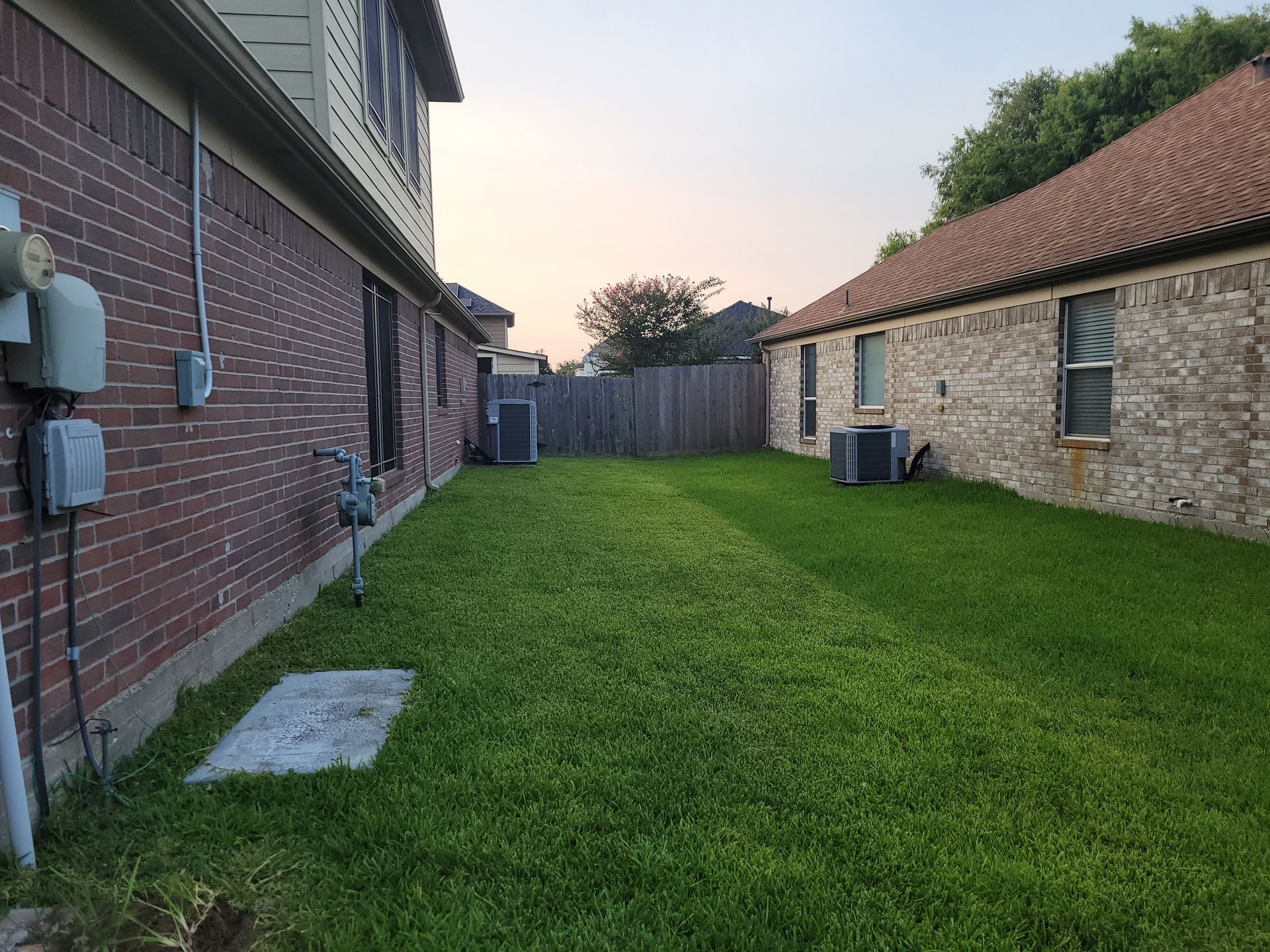 Green backyard between two brick houses, with air conditioning units and a wooden fence.