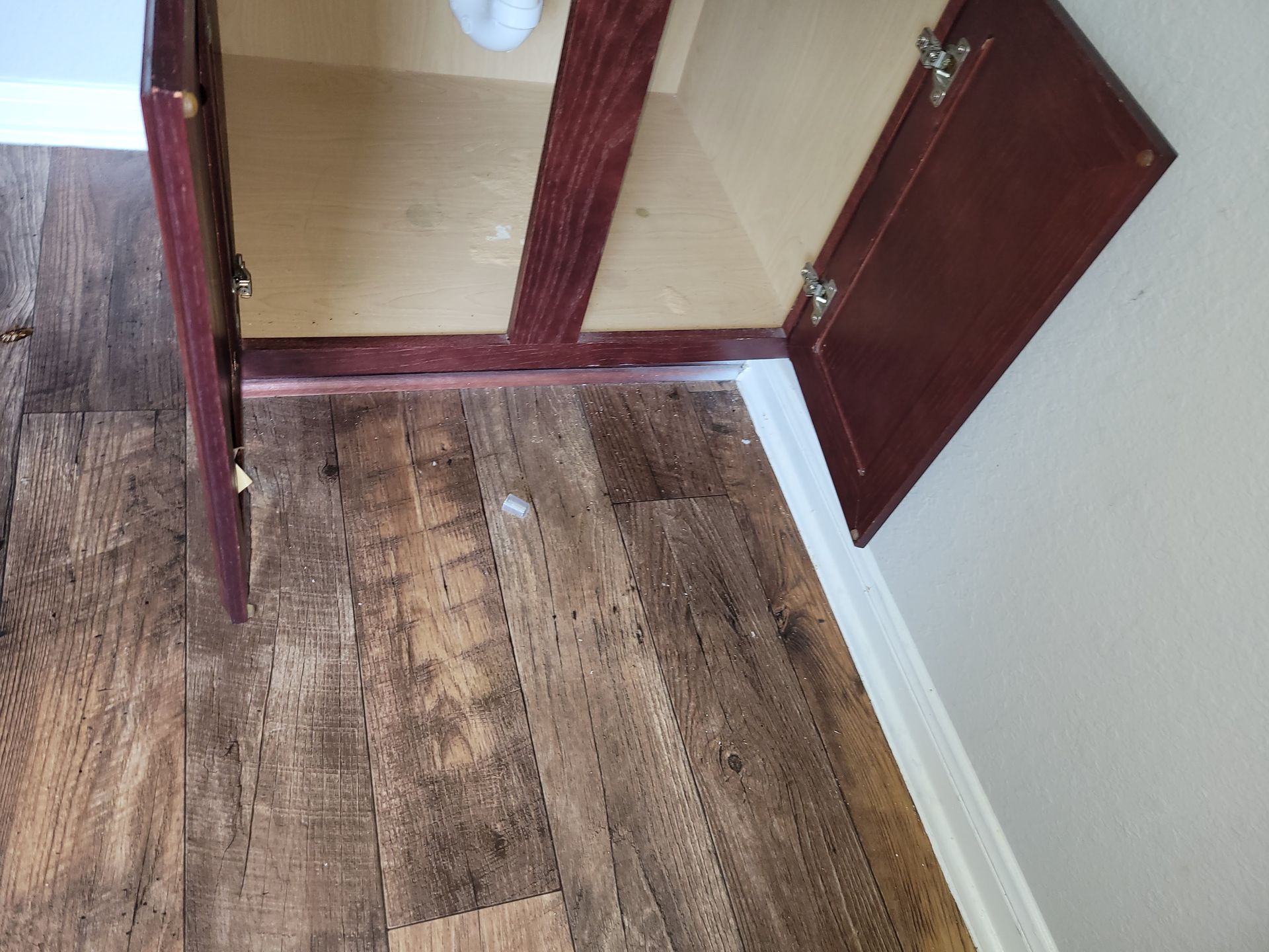 Open bathroom cabinet doors, dark wood, installed on wood-look flooring next to white baseboard.
