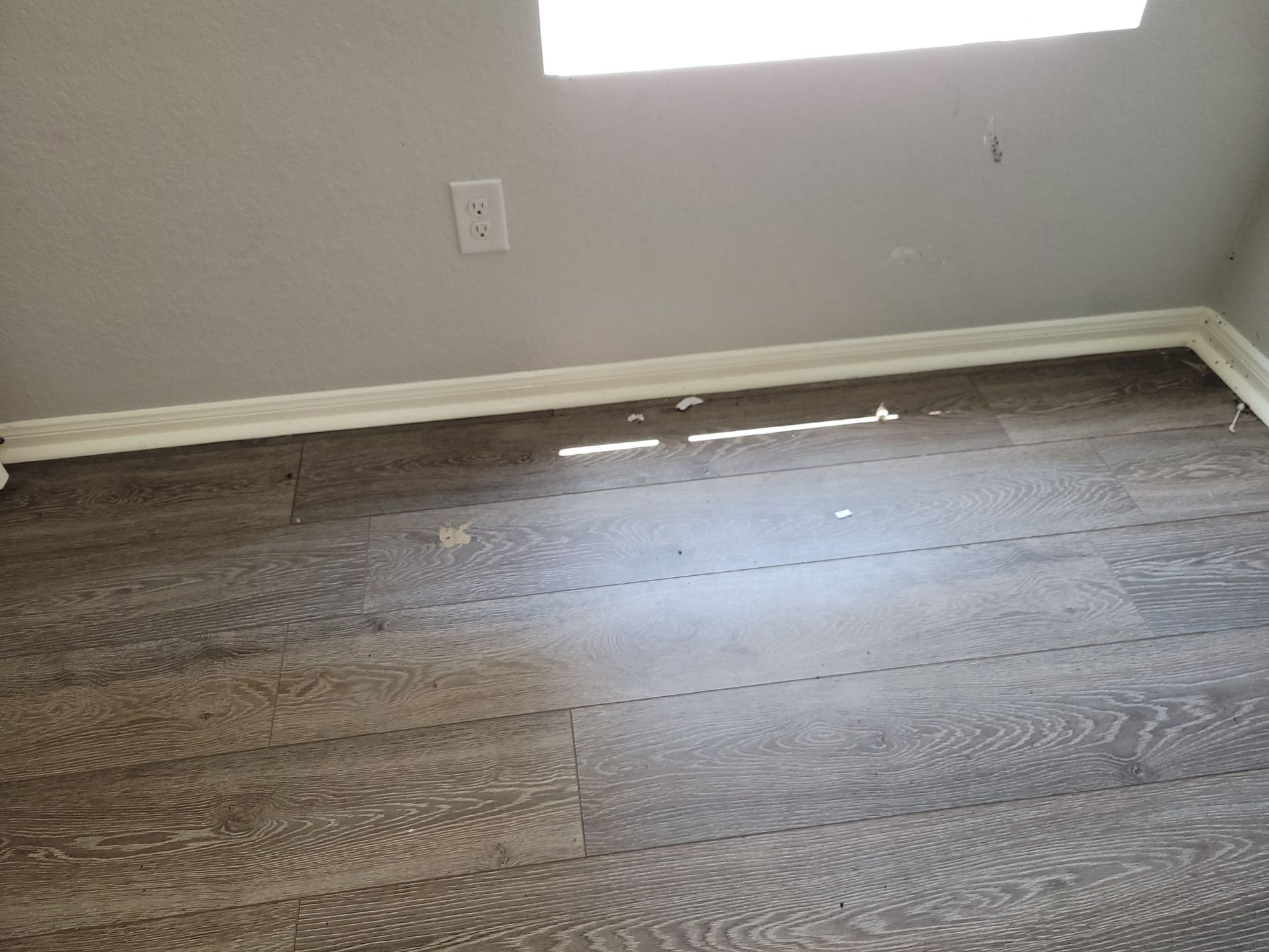 Wood floor with damaged trim and a power outlet near a window.