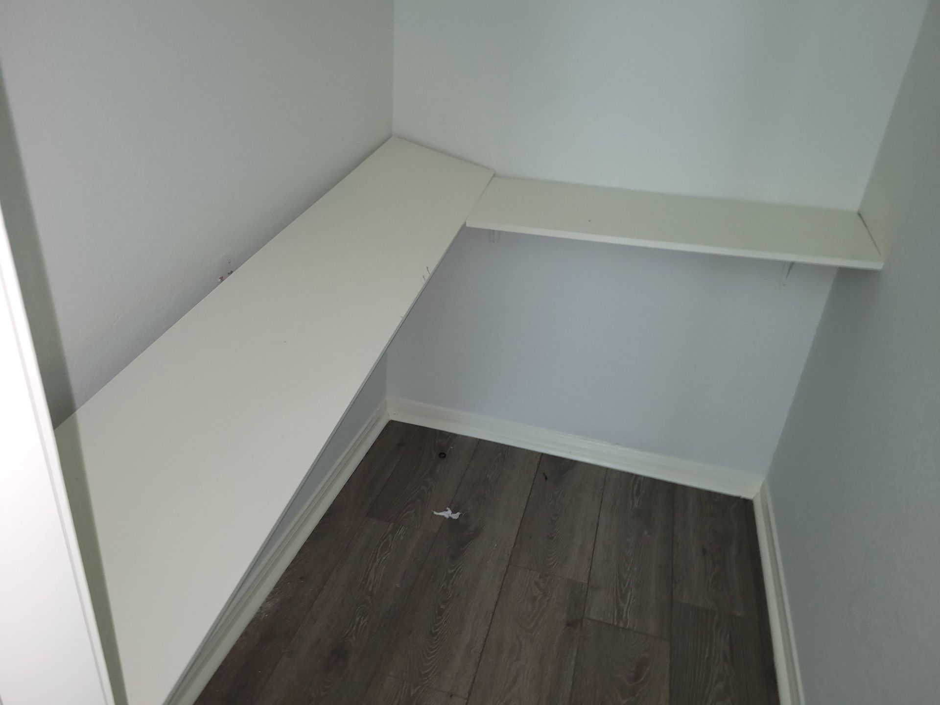 White shelves in a corner closet with dark wood-look flooring and light gray walls.
