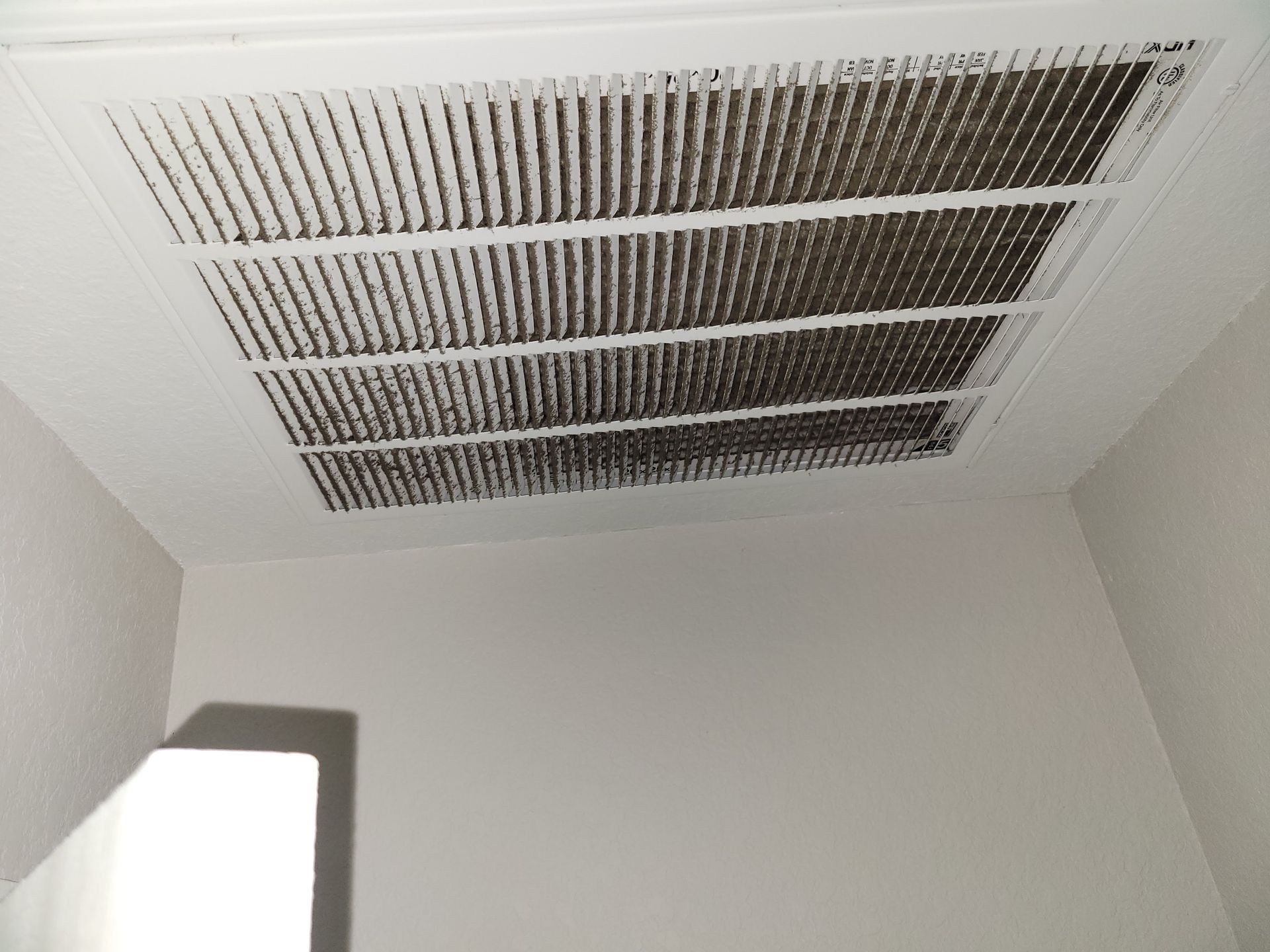 Ceiling vent covered in dark dust, in a corner of a white-walled room.