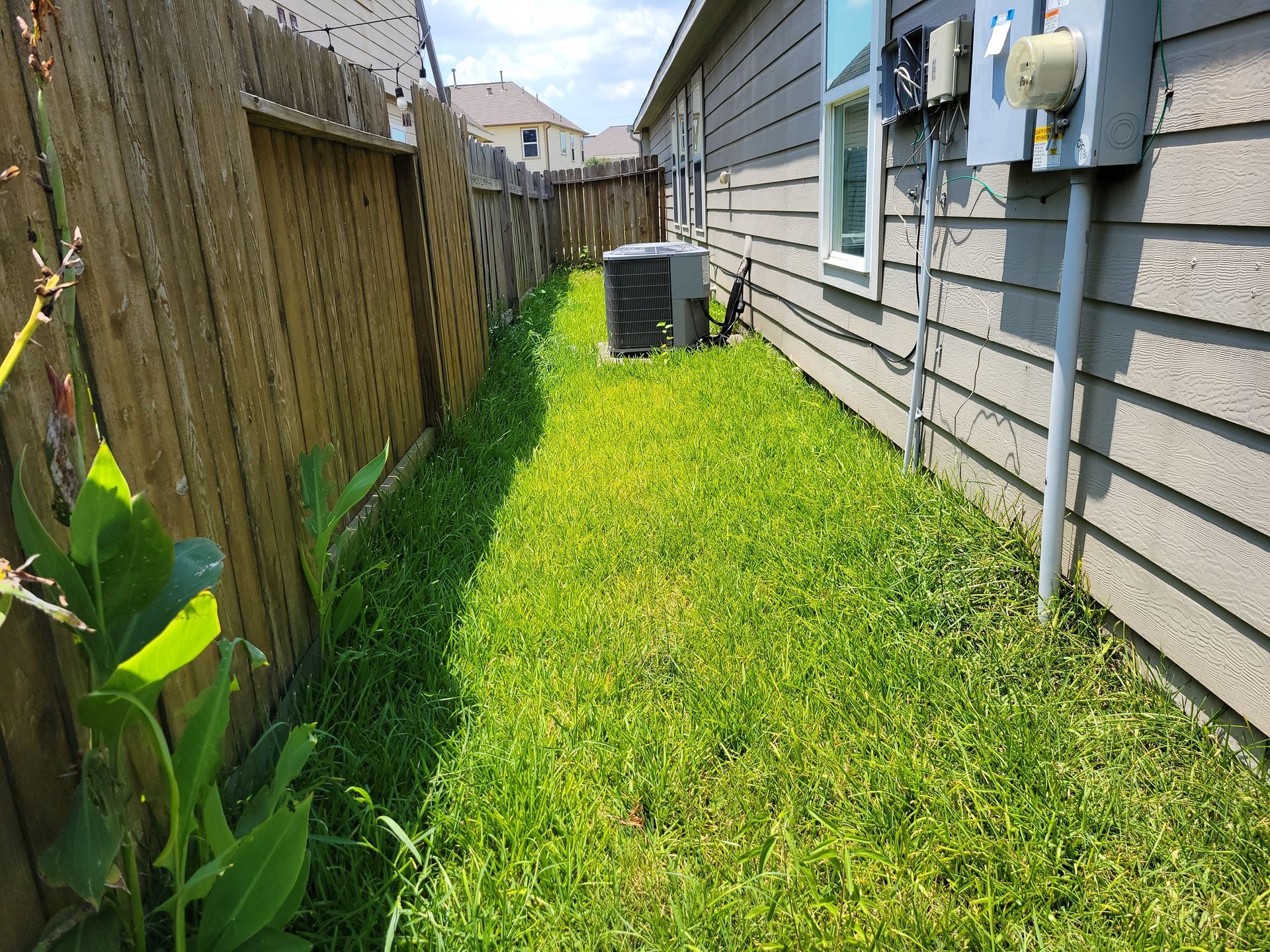 Narrow grassy yard between wooden fence and house with AC unit.
