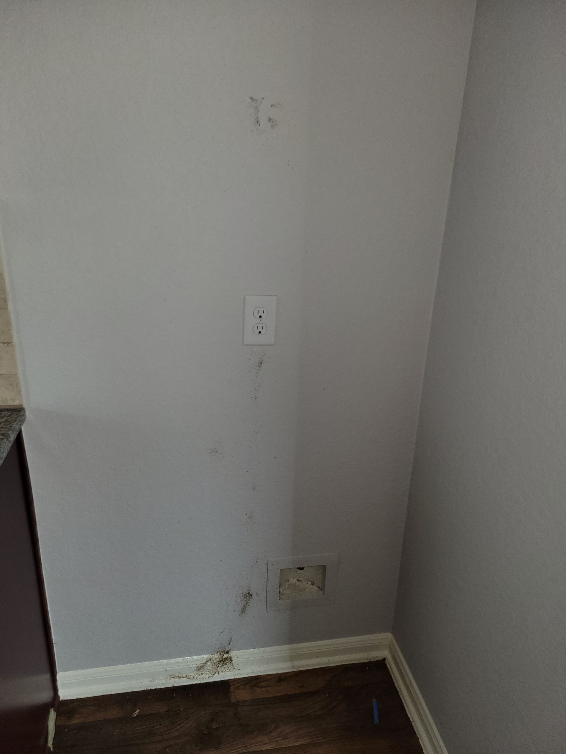 Gray wall with outlet, water damage, and a hole near the floor, in a corner.