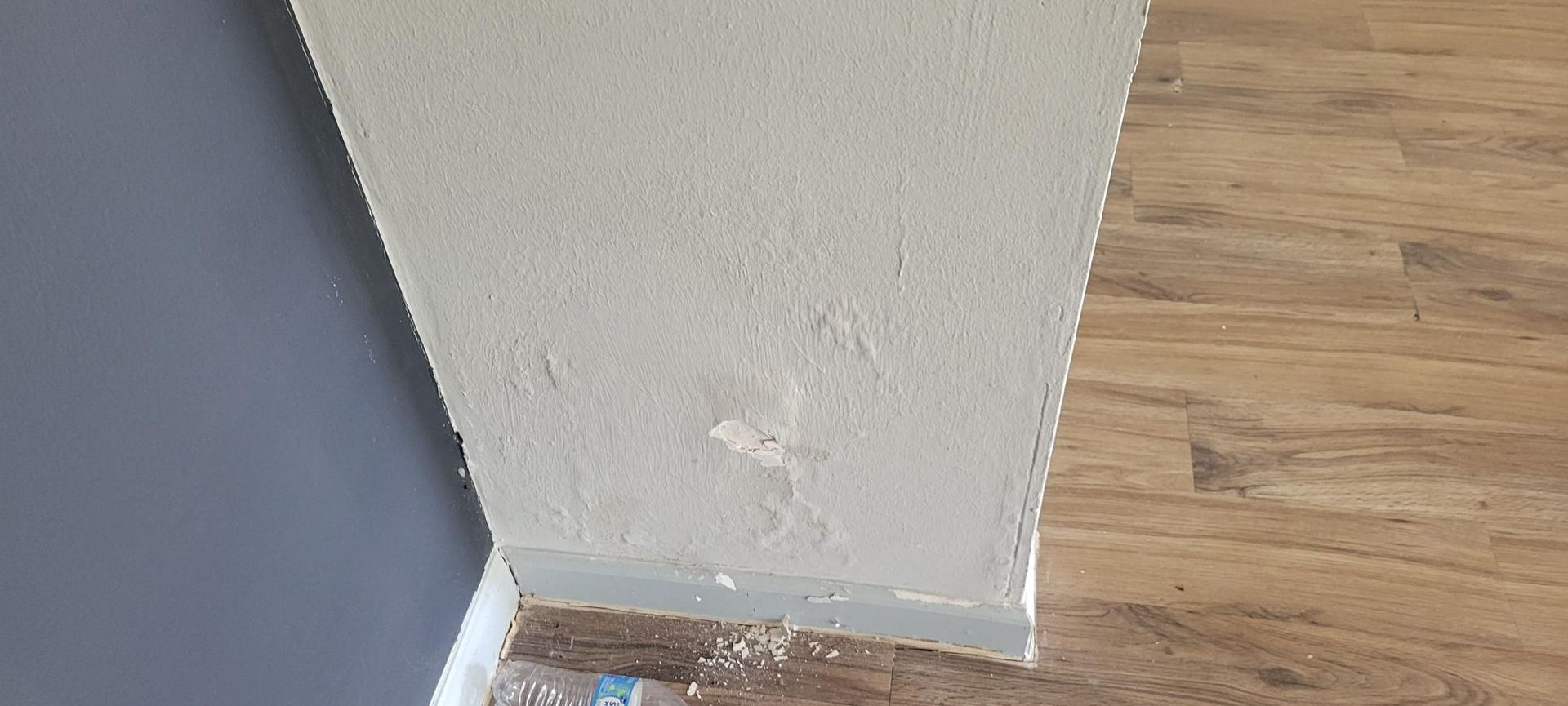 Peeling paint on a light-colored wall, next to a gray wall and brown wood-look flooring.