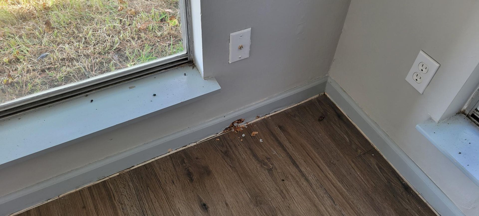 Corner of a room with damage to the baseboard near a window and electrical outlet.