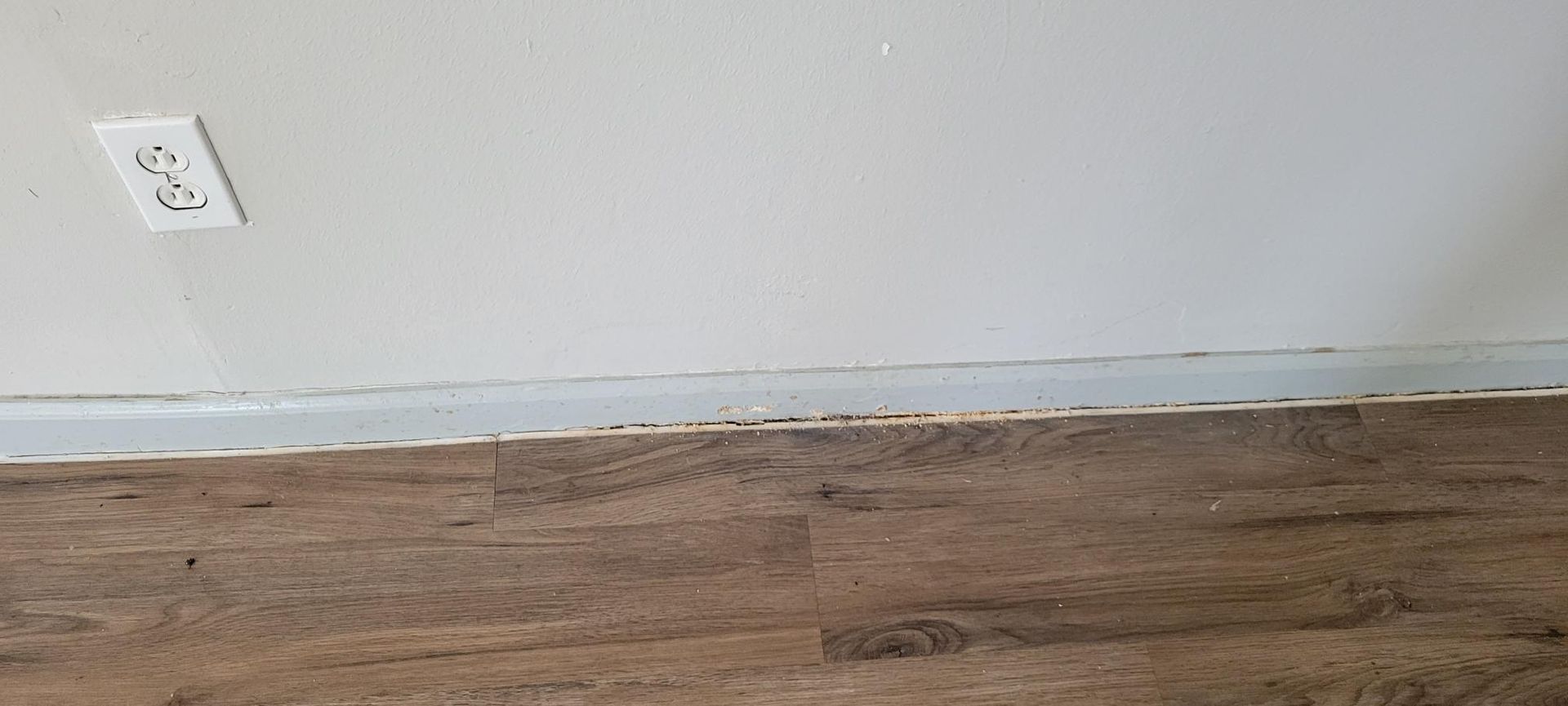White wall, outlet, and wood flooring. A gap between the floor and wall is visible.