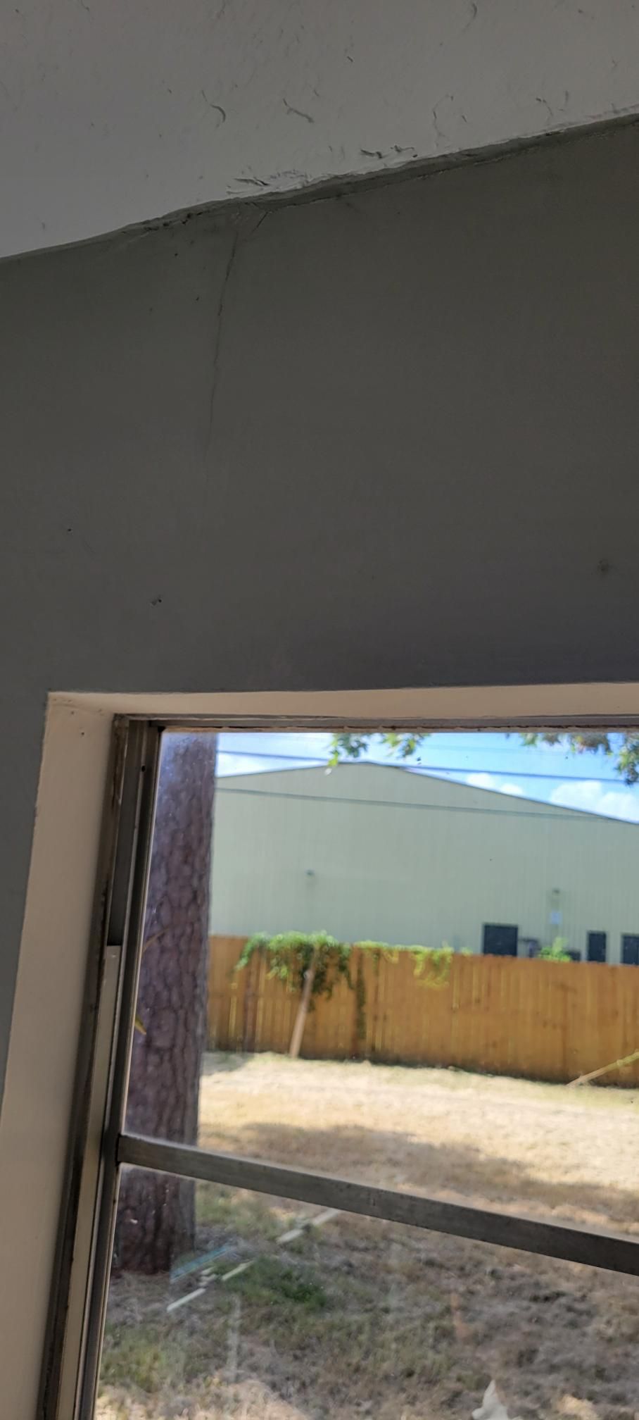 View through a window, showing a backyard with brown grass, wooden fence, and a white building.
