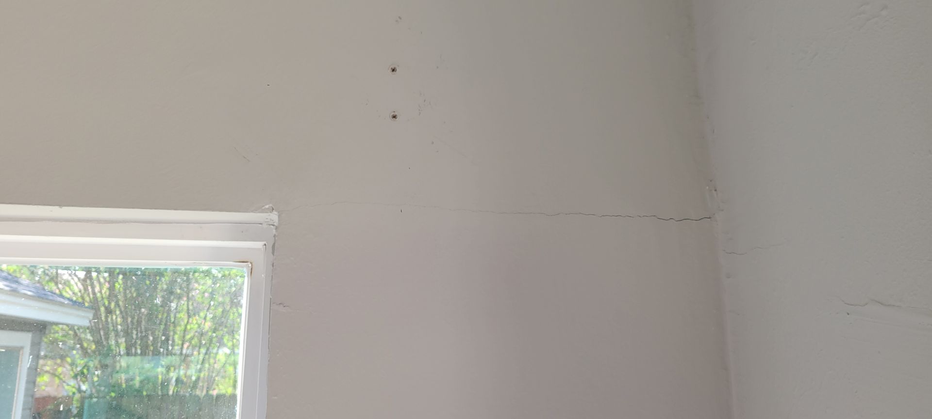 Interior corner of a room with a window showing greenery, featuring a white wall with some blemishes.