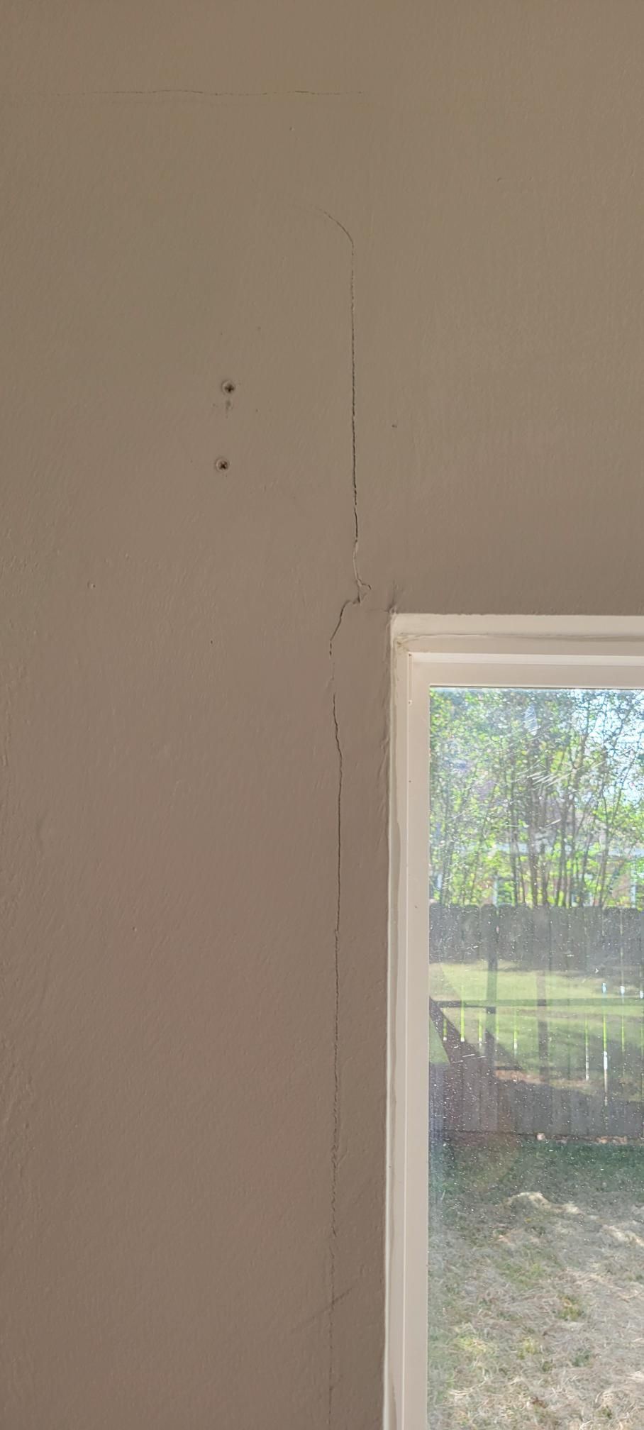 A light beige interior wall with a vertical crack, next to a window with an outdoor view of trees and grass.