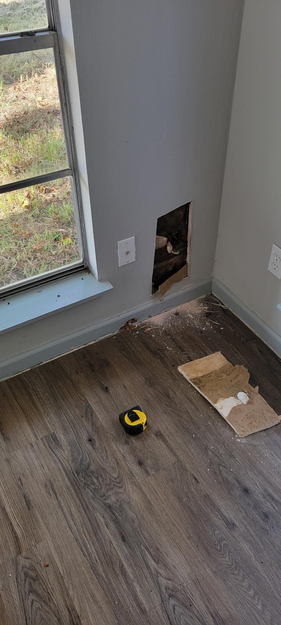 A hole in a grey wall, next to a window, with debris and a tape measure on the wood floor.