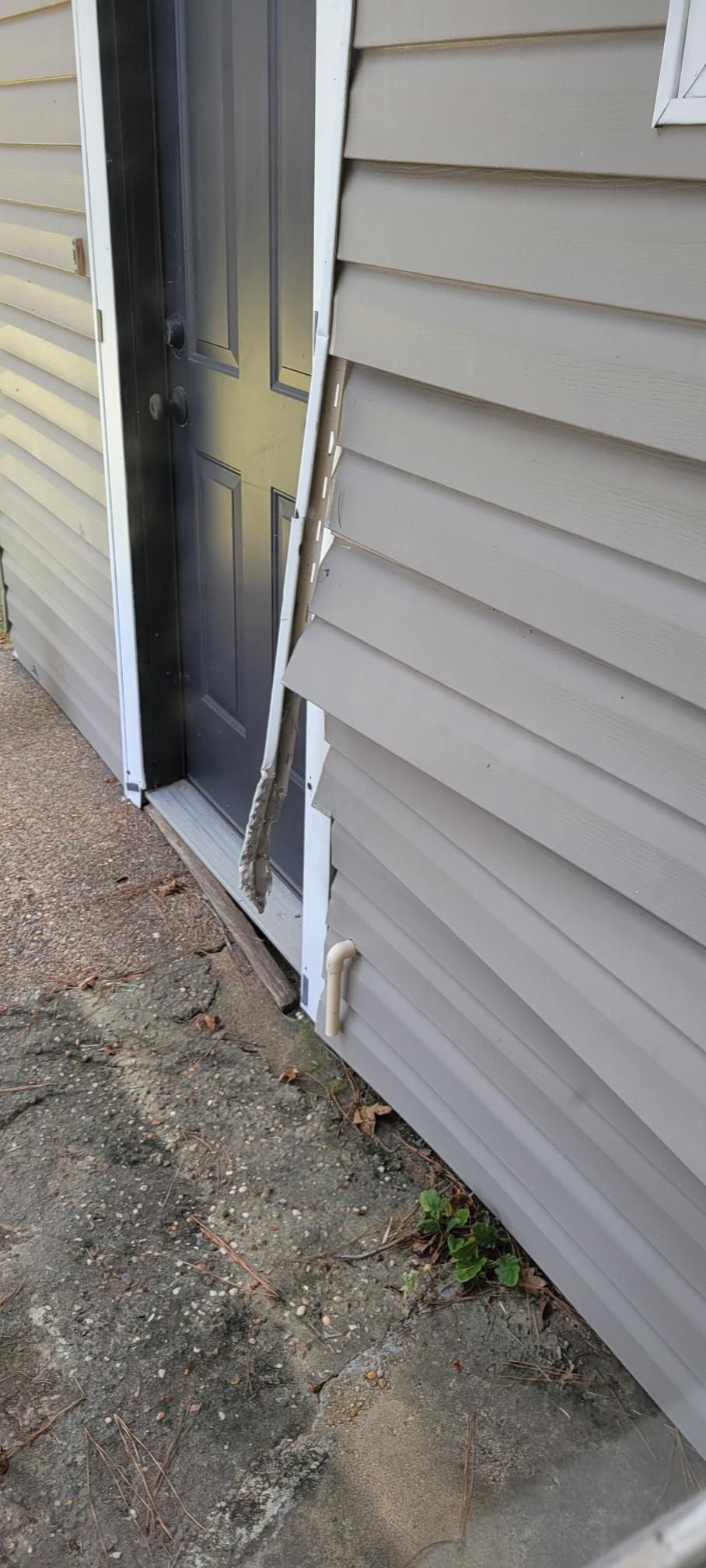 Damaged siding around a gray door. White trim is peeling away. Cracked concrete and weeds nearby.