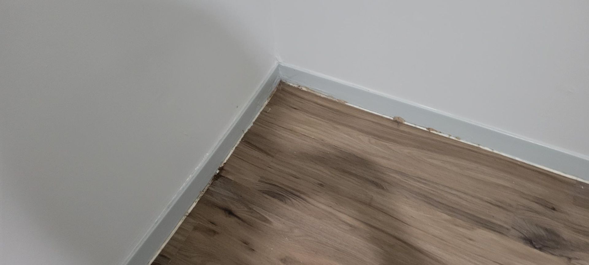 Corner of a room with white wall and wooden floor. A white baseboard runs between the two.