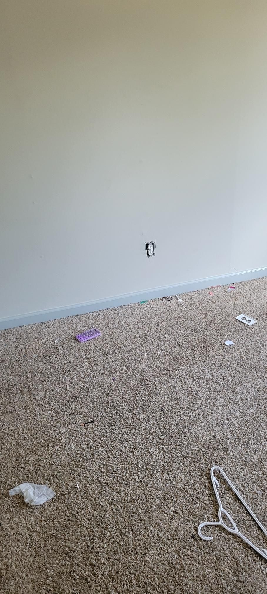 Beige wall, electrical outlet, and carpeted floor with scattered debris, and hangers.