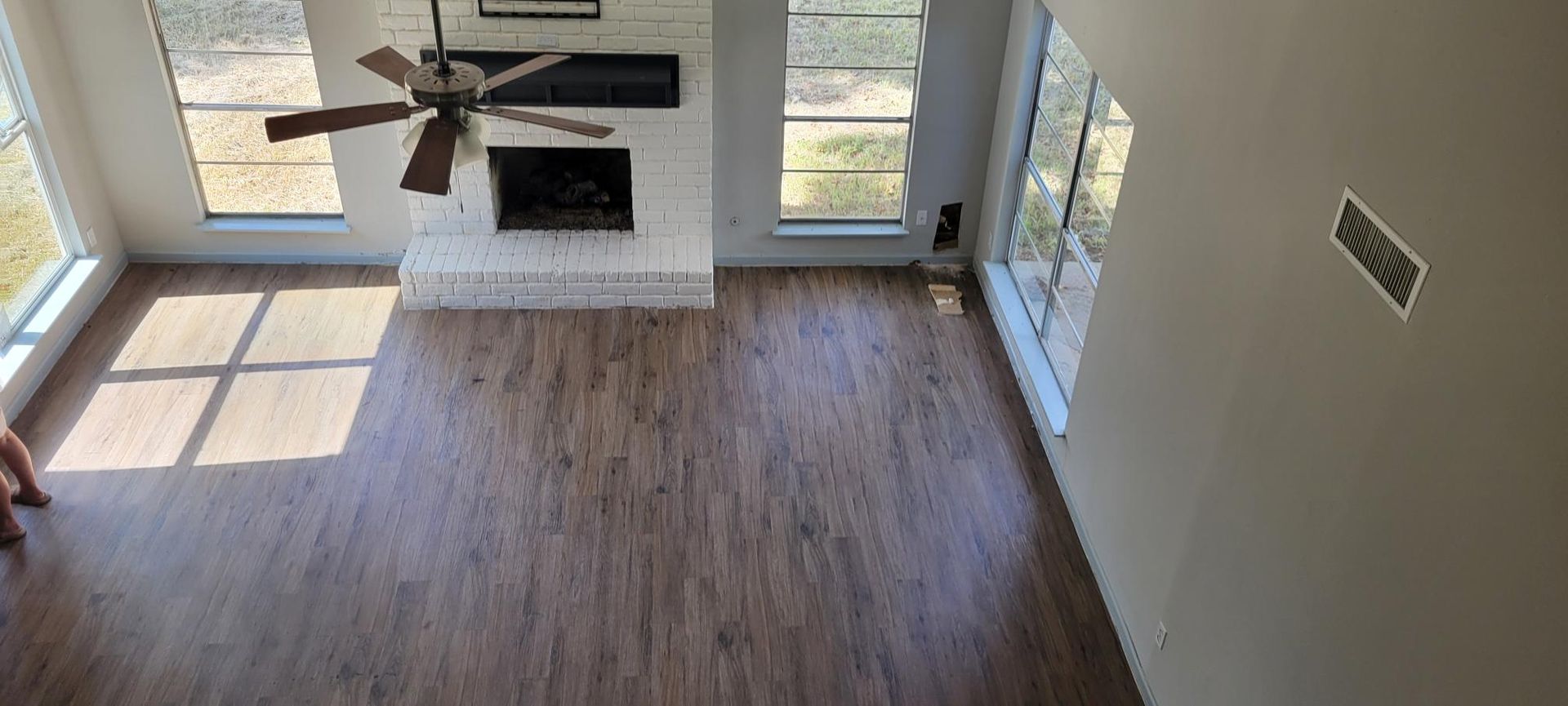 An overhead shot of a living room with wooden flooring, a brick fireplace, large windows, and a ceiling fan.