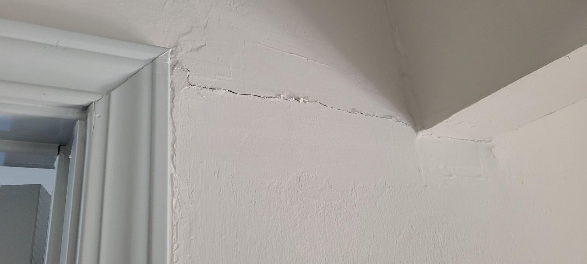White wall with a long horizontal crack, near the corner of a door frame and ceiling.