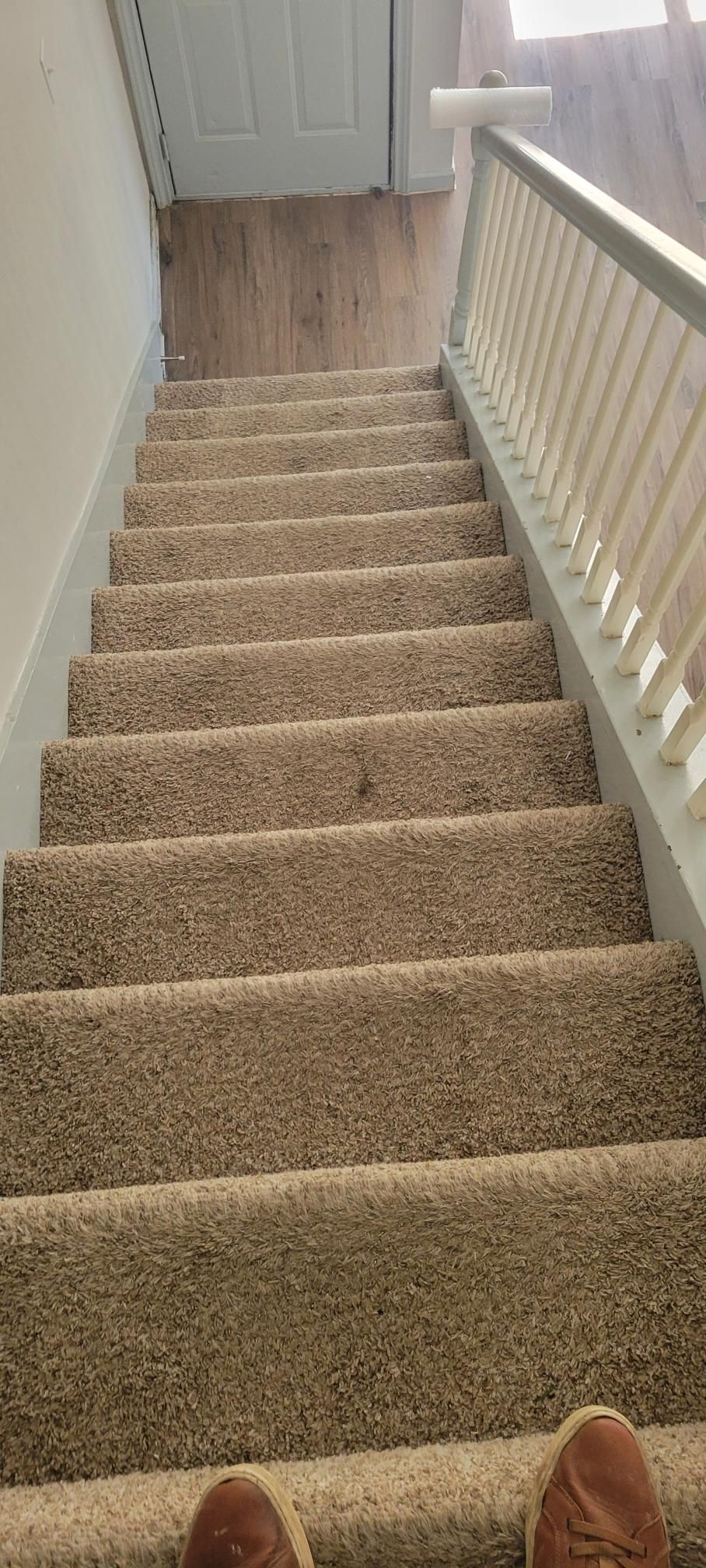 Staircase with carpet steps and white railing, viewed from above, with the viewer's feet in the foreground.
