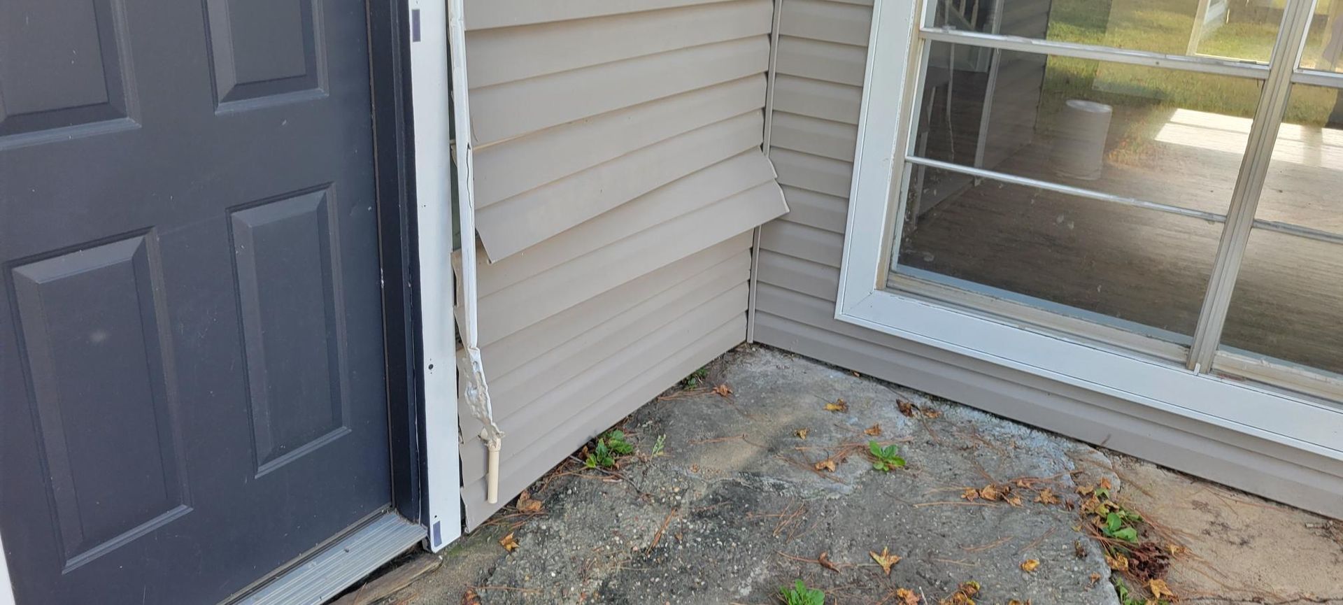 Exterior corner of a building, showing door, window, and damaged siding. Gray door, tan siding, concrete surface.