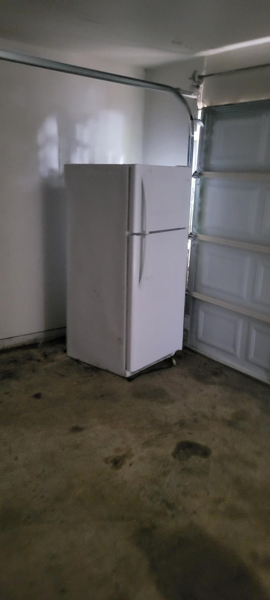 White refrigerator in a garage setting, floor stained. Garage door partially open.