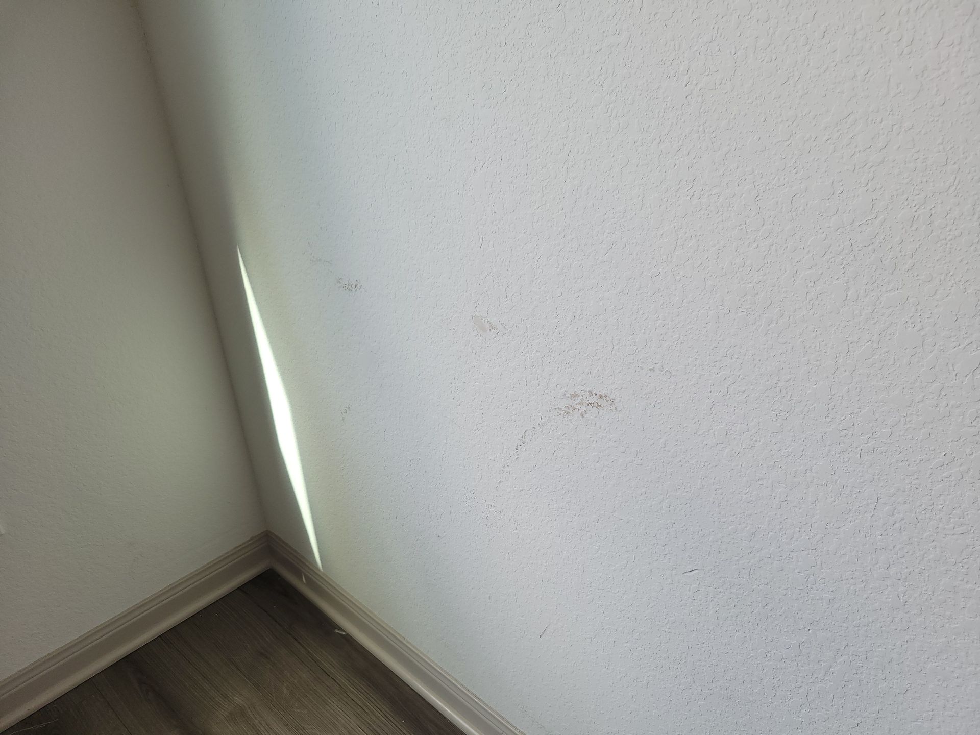 Corner of white speckled wall with light shining from the side; wood floor visible.