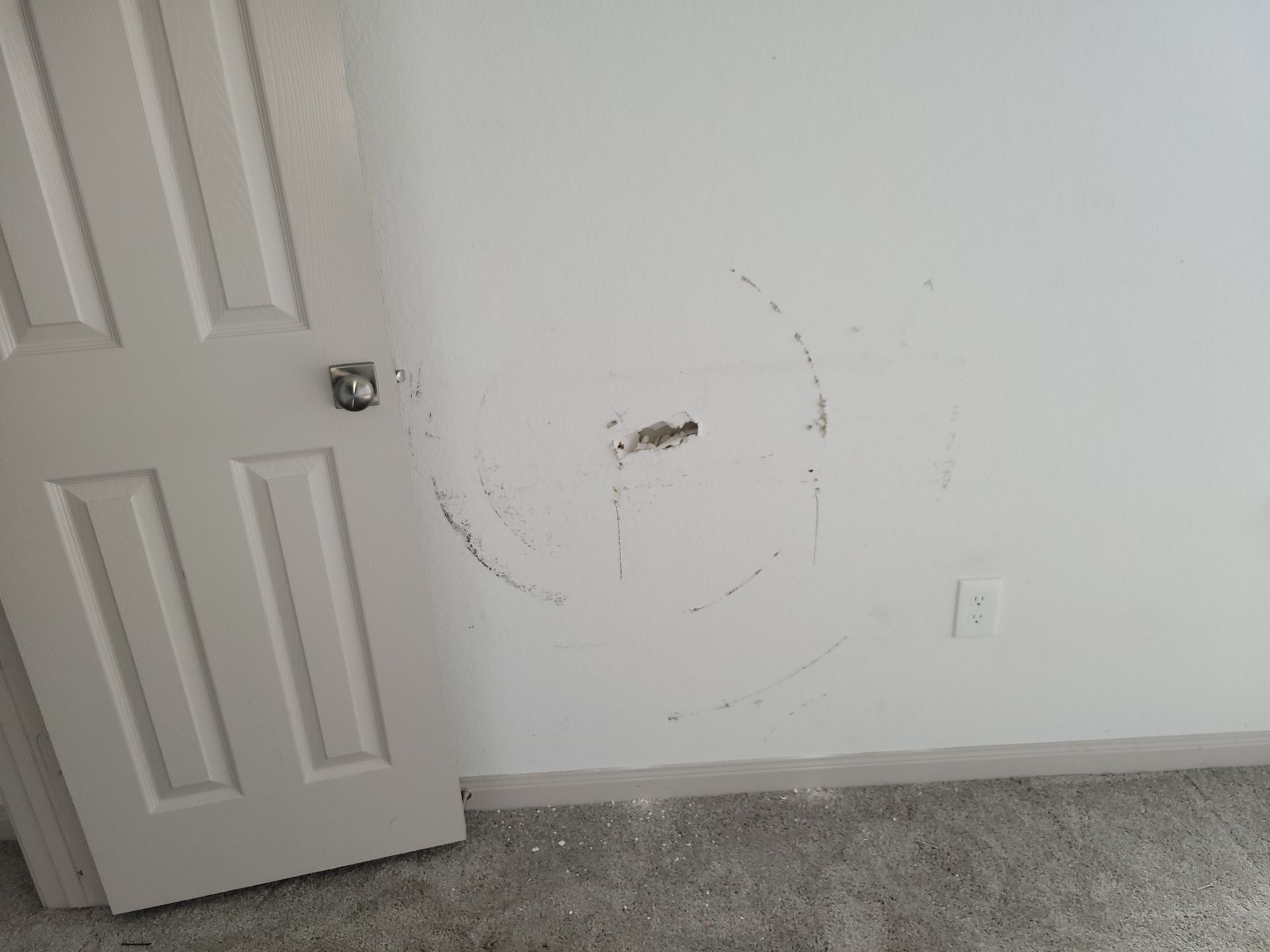 White wall with circular damage and a hole near the center, next to a closed white door.
