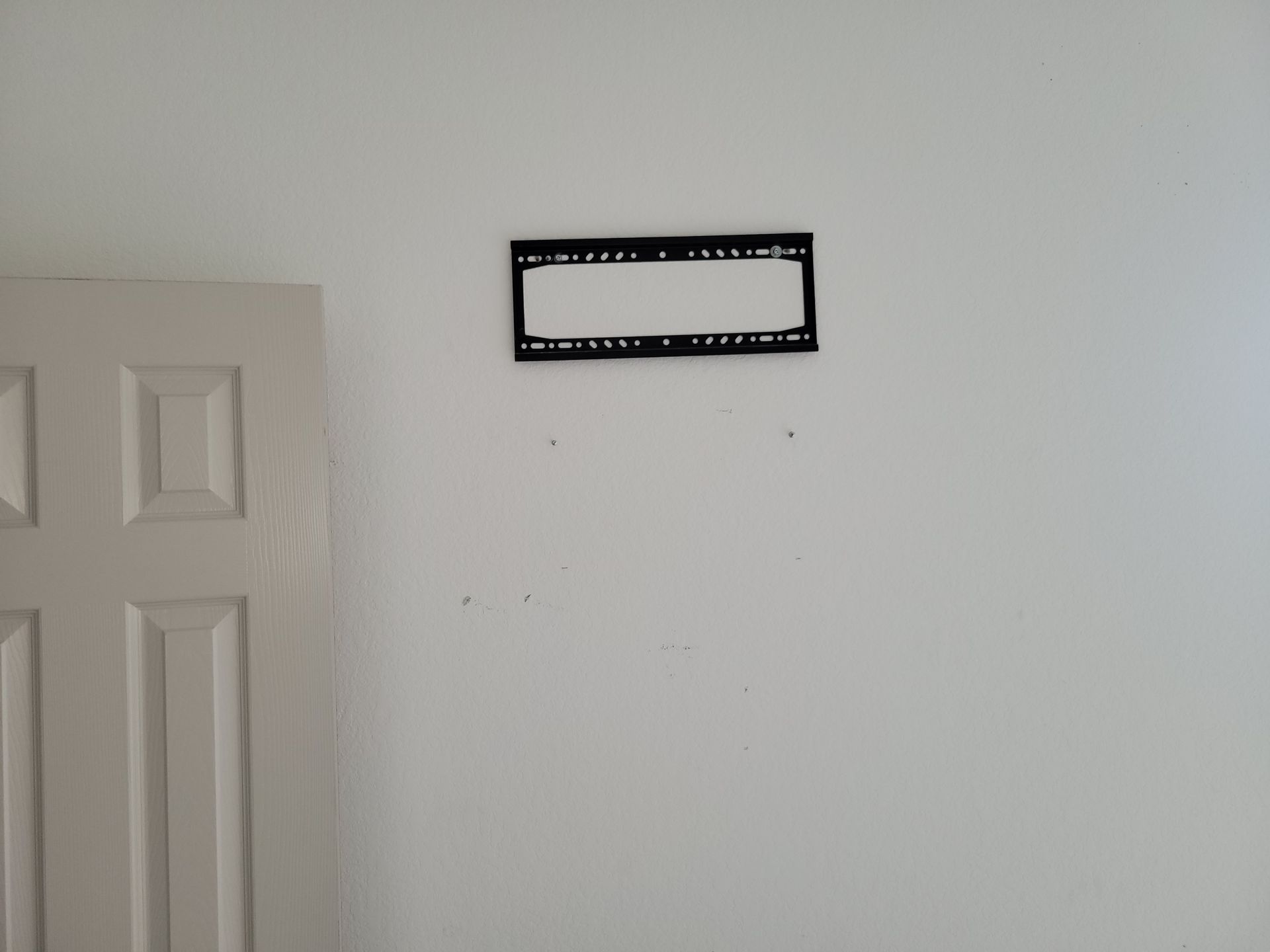 White wall with a mounted black TV bracket and some screw holes. A white door is on the left.
