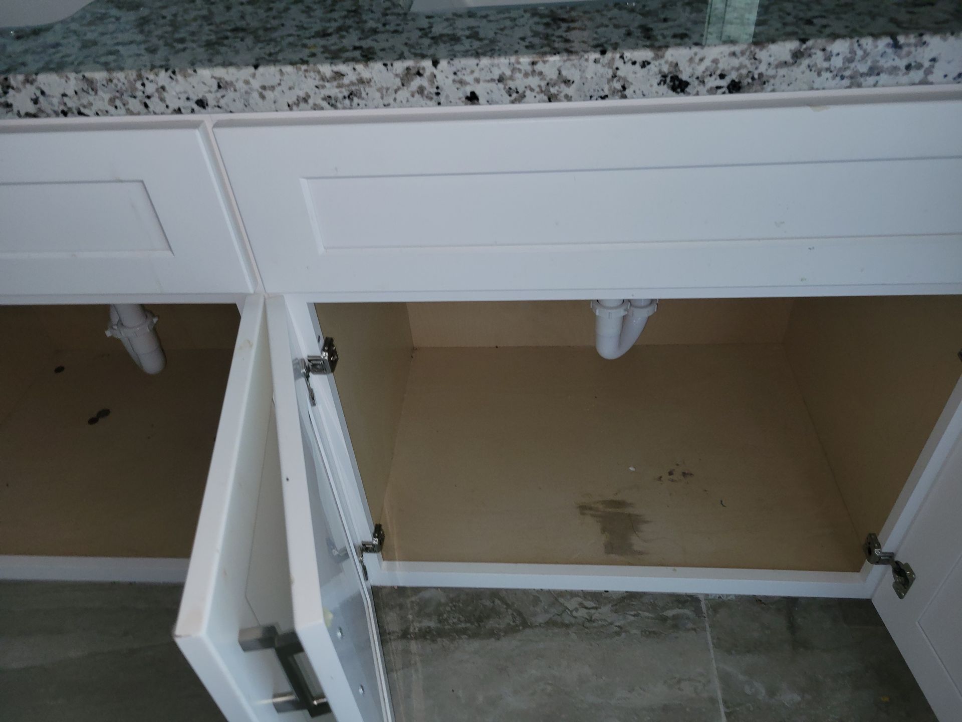 Open white bathroom cabinet doors, interior view, beige and white, plumbing visible.
